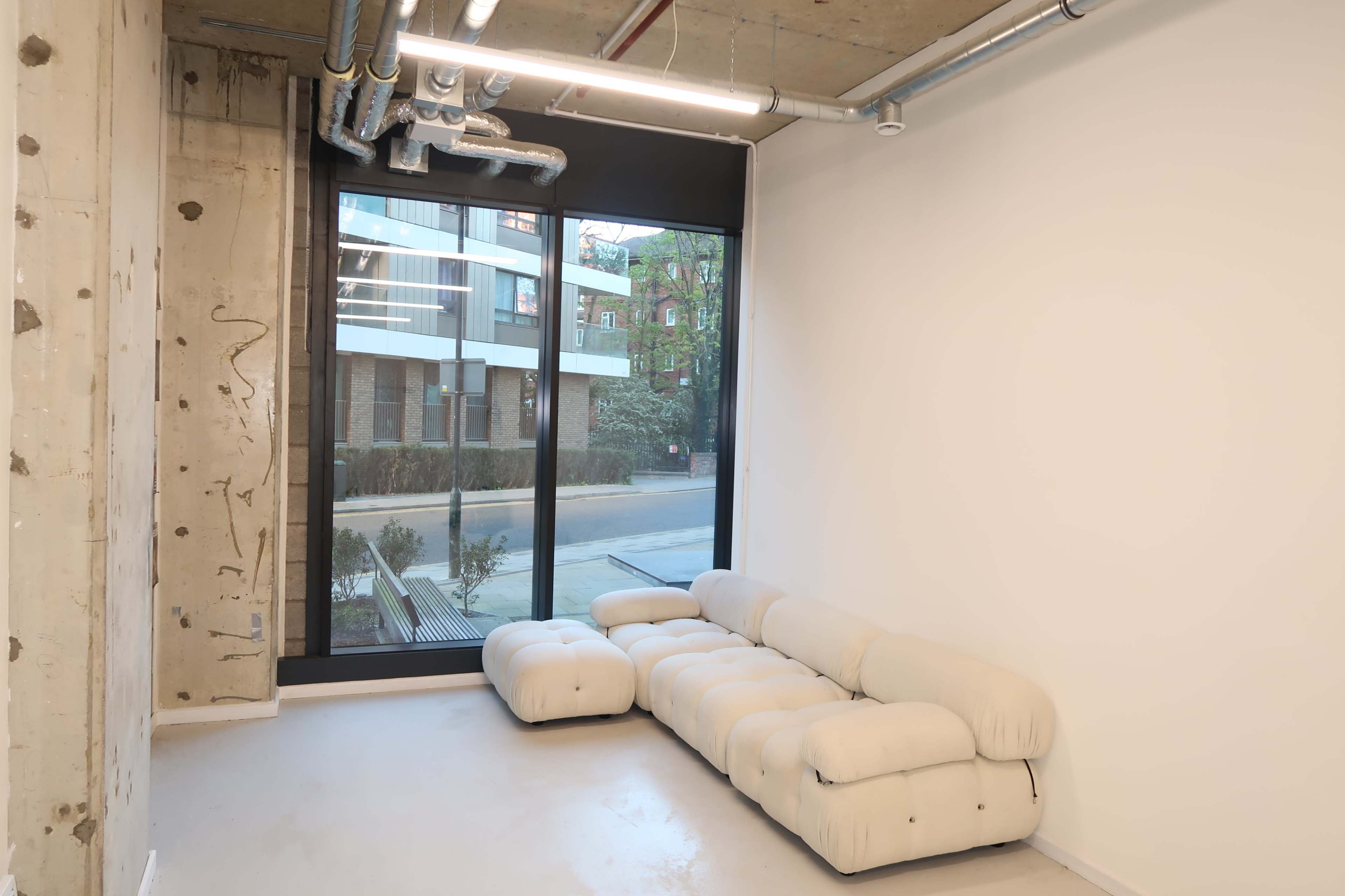 A light-colored, modular sofa is positioned against a wall in a room with large windows overlooking a street and buildings outside.
