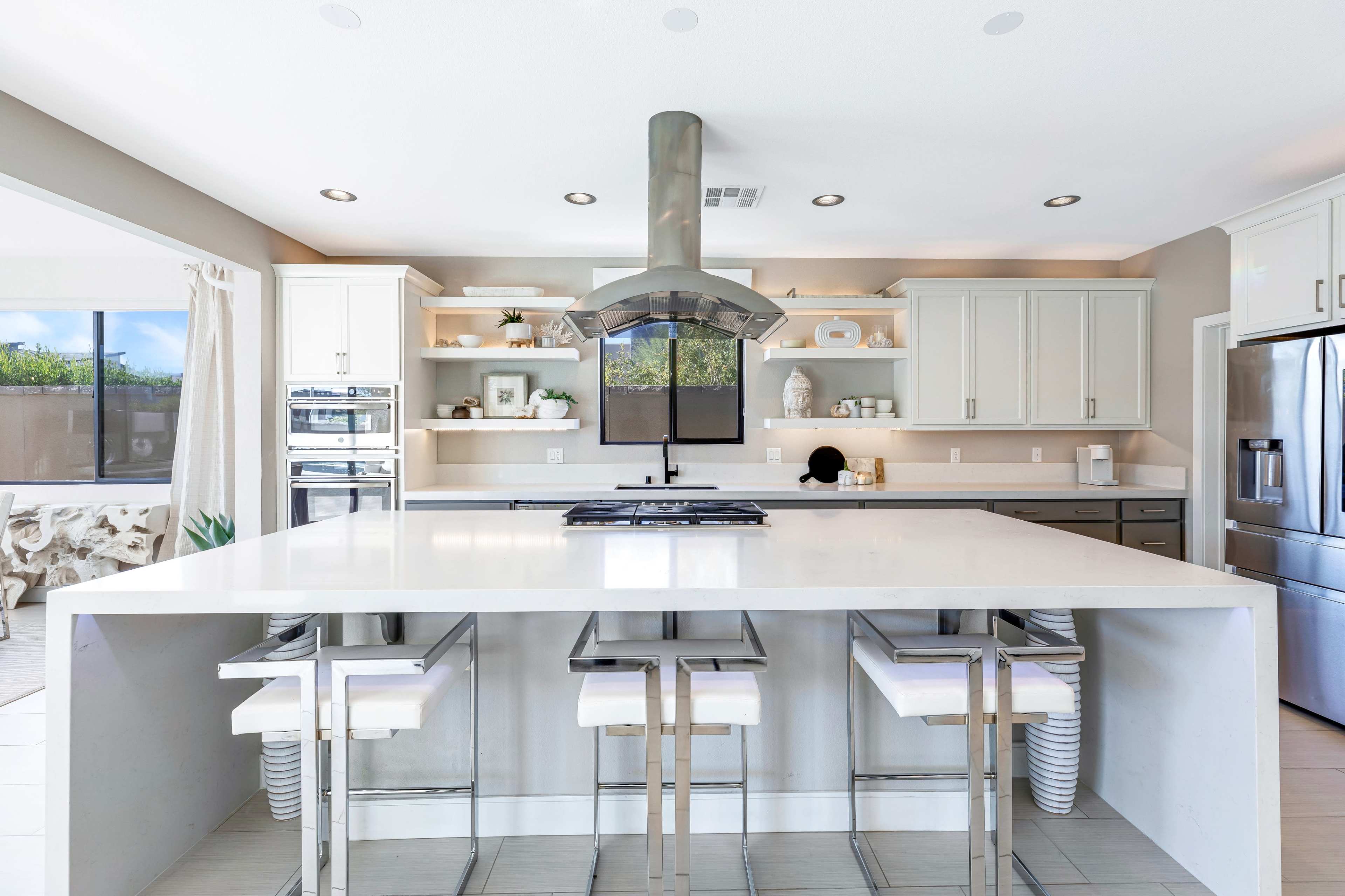 A modern kitchen features a central island with bar stools, a cooktop, stainless steel appliances, and open shelves displaying various kitchen items.