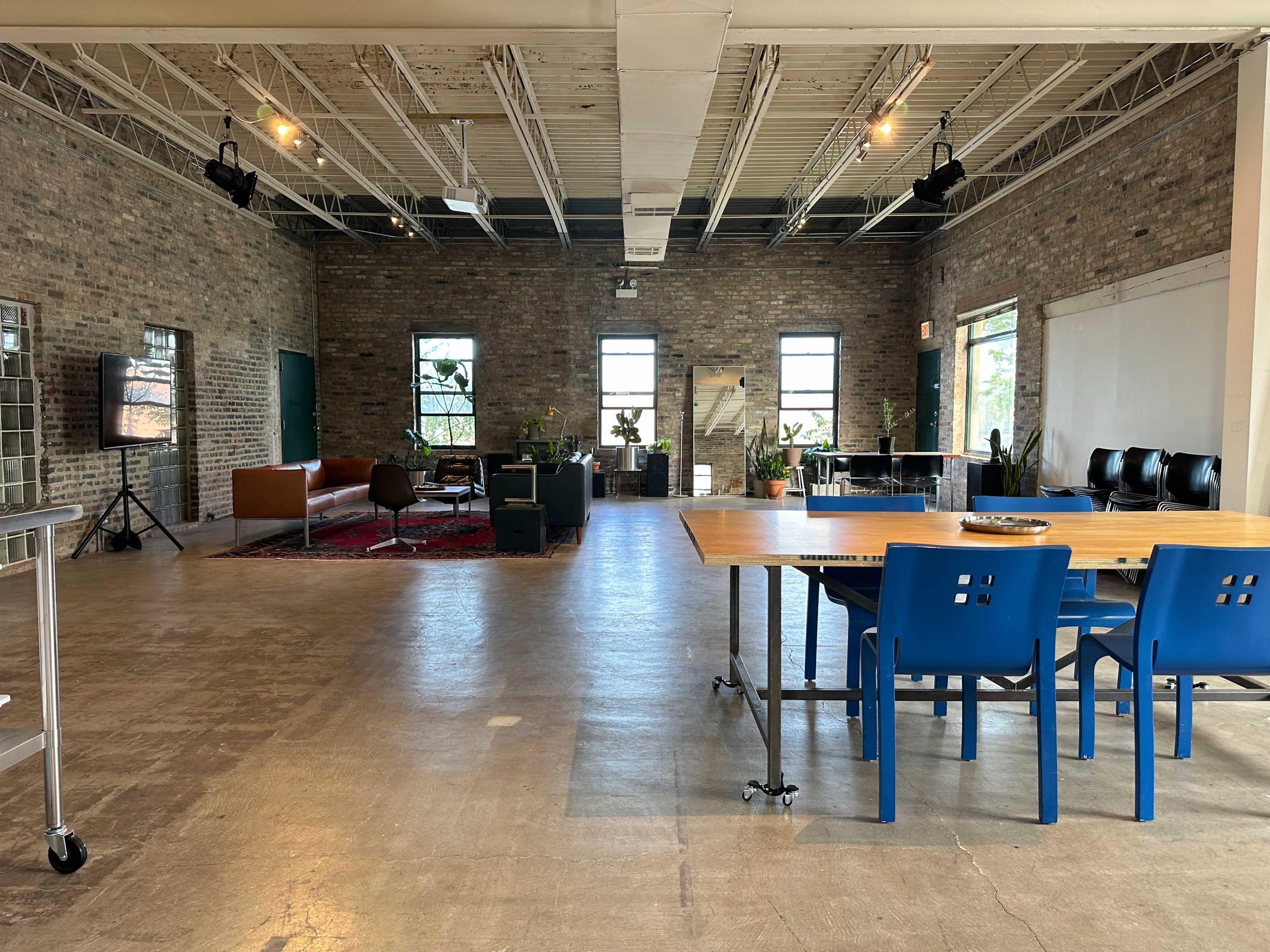 The image shows a spacious room with exposed brick walls, a wooden dining table with blue chairs, and a seating area featuring a sofa and plants.