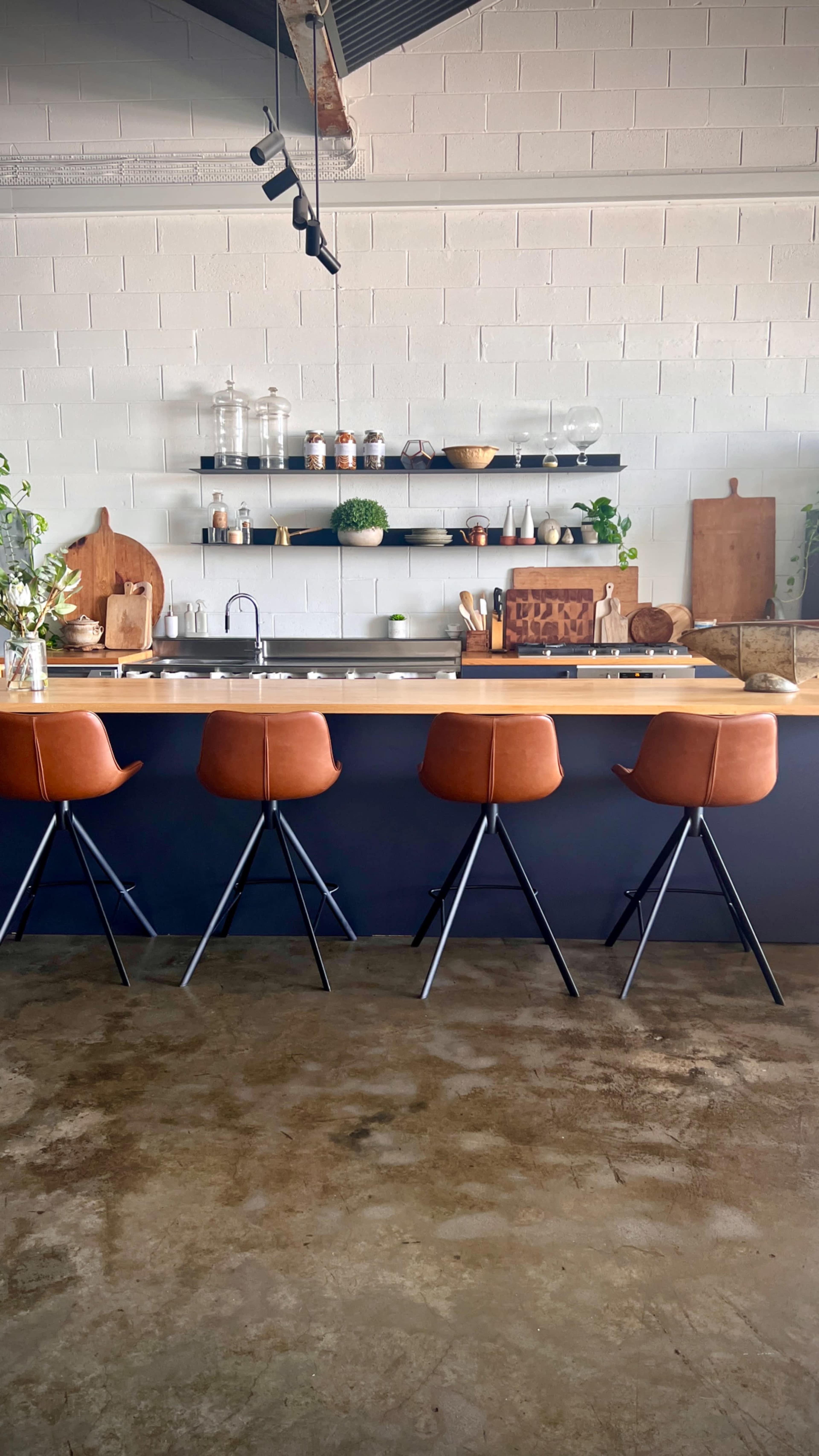 The image shows a modern kitchen with a long, wooden countertop, four brown leather bar stools, and open shelves displaying various kitchenware and plants.