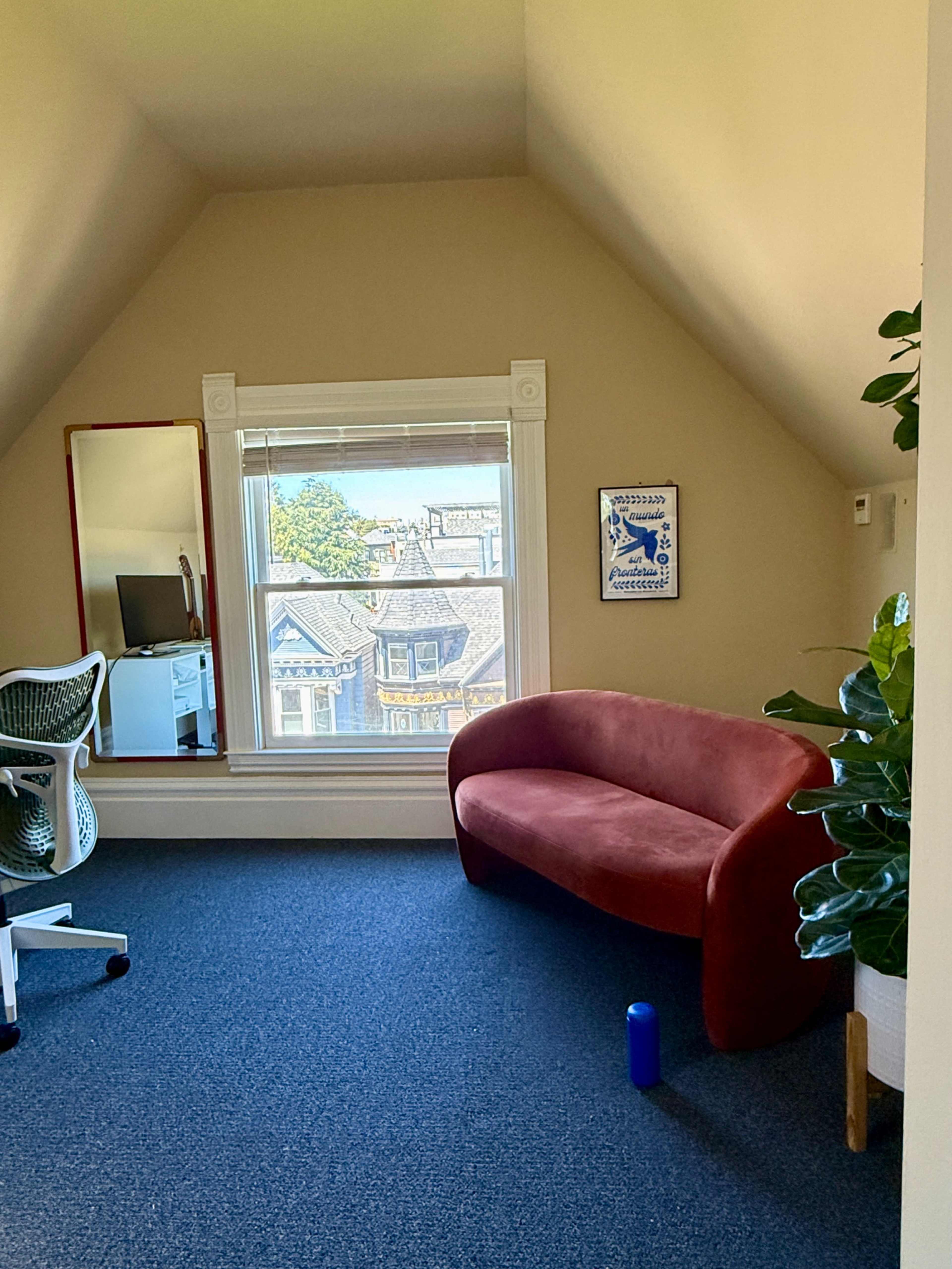 The image shows a cozy office space featuring a red curved sofa, an office chair, a desk with a computer, and greenery near a window with a view of neighboring houses.
