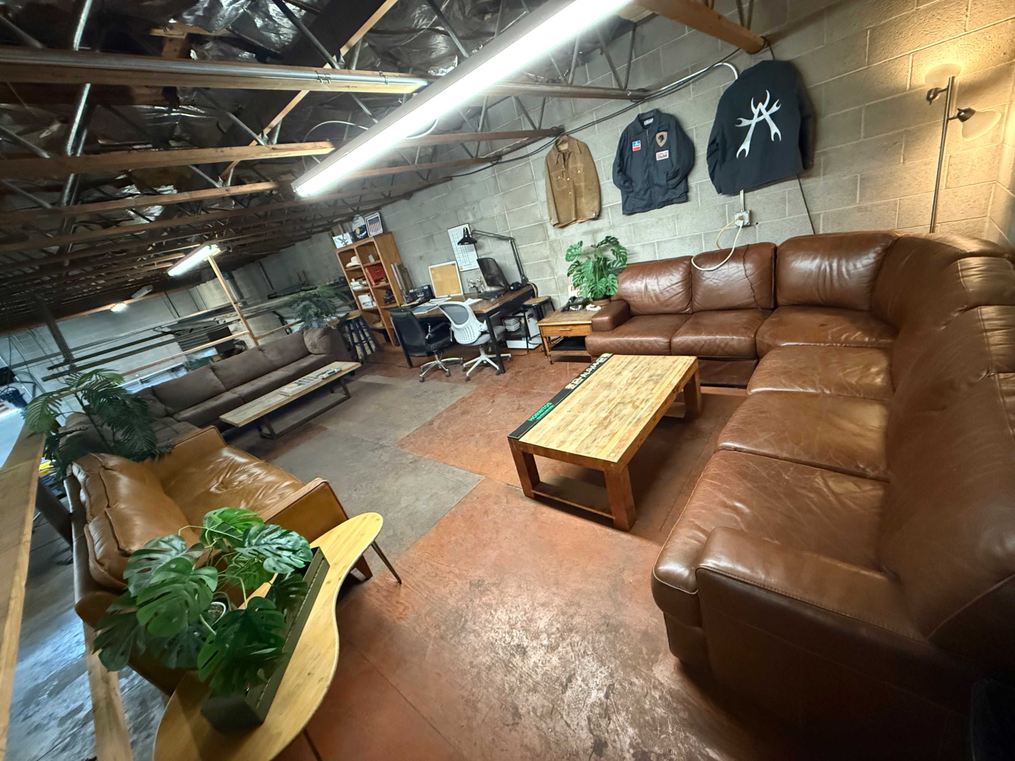 The image shows a spacious, well-lit room with leather couches, a wooden coffee table, and an office area featuring a desk and chair.
