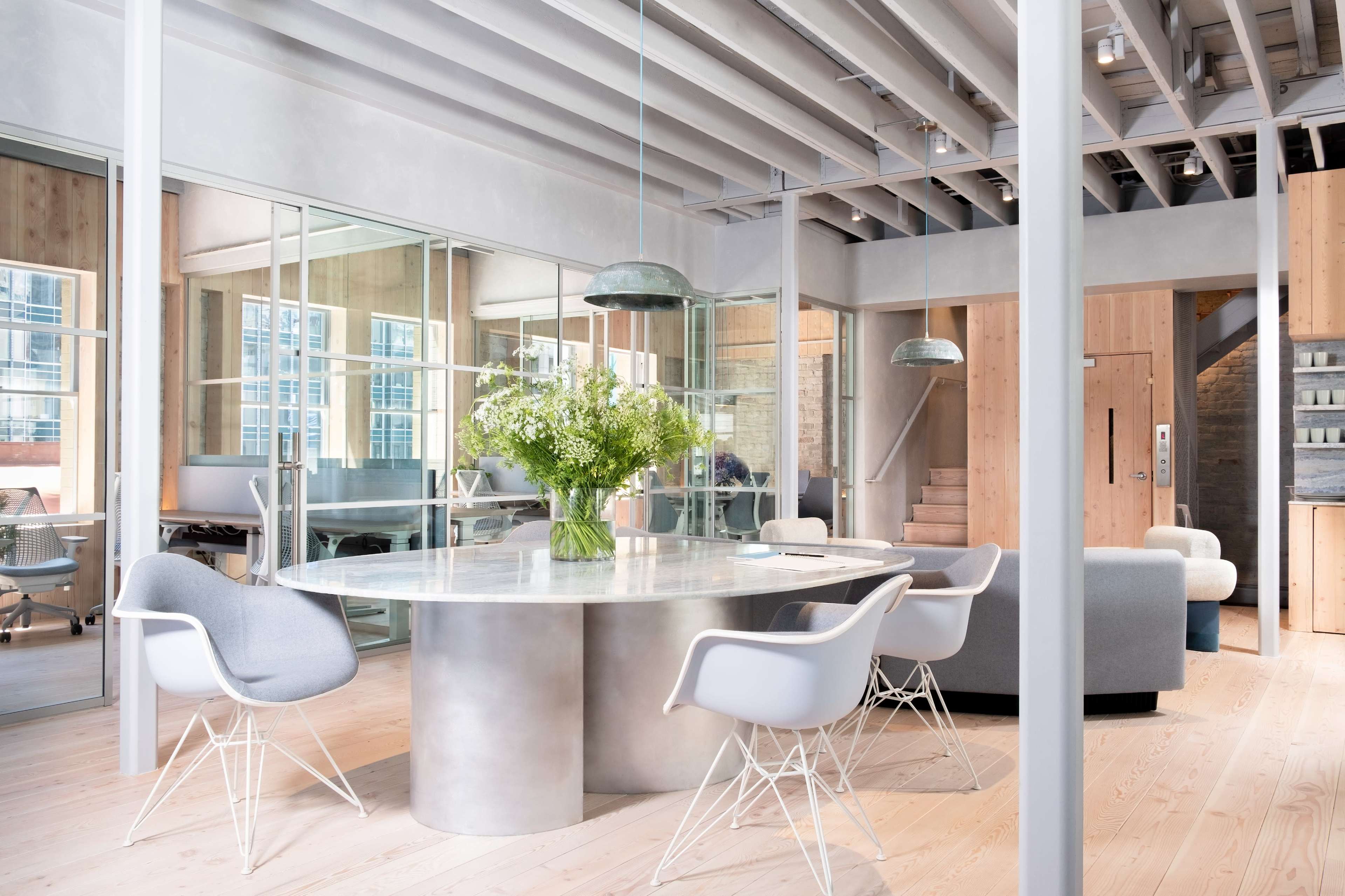 The image shows a modern office space featuring stainless steel and glass elements, a round table with a floral centerpiece, and minimalist white chairs arranged around it.