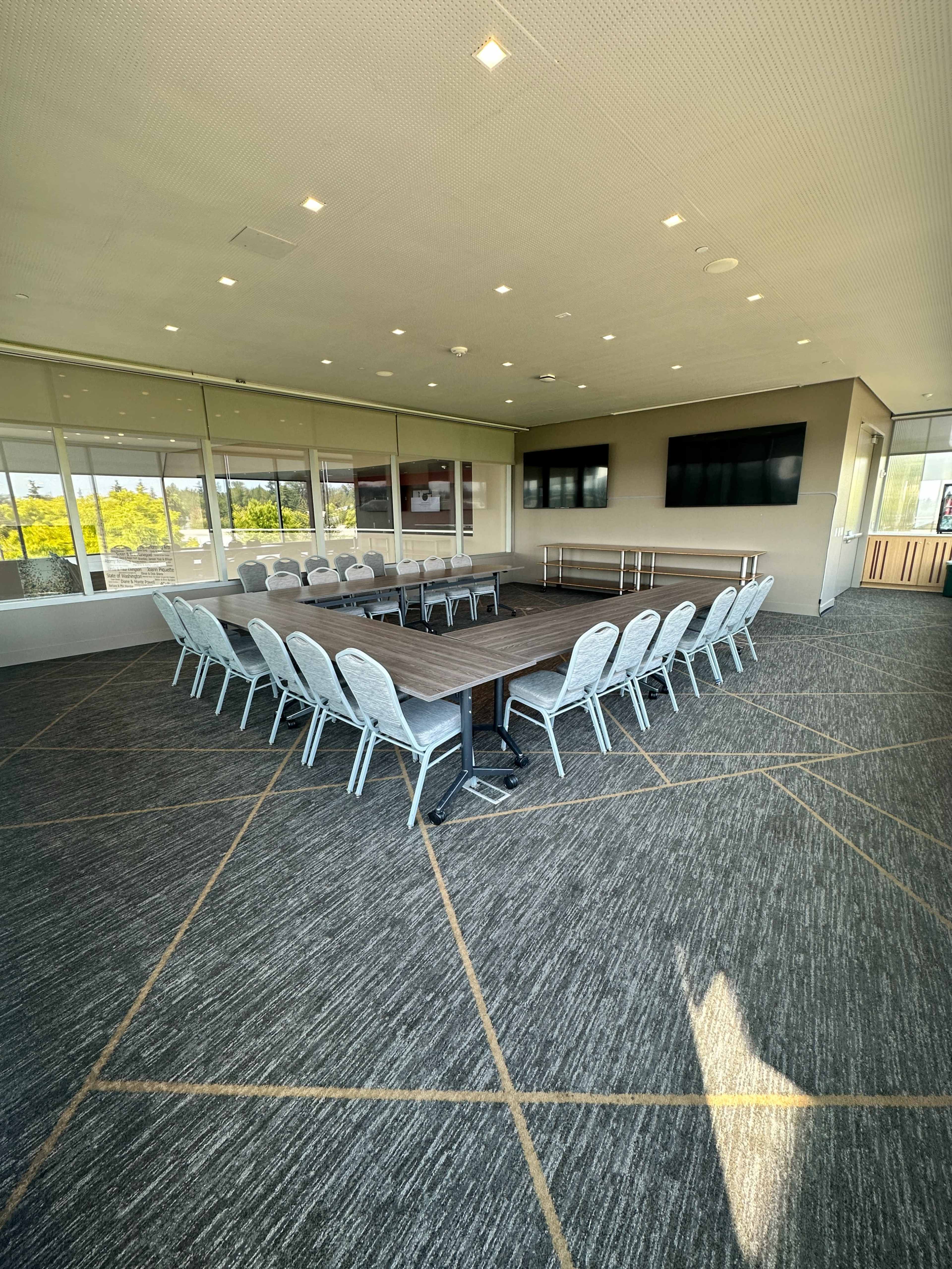 A spacious conference room features a large rectangular table surrounded by chairs, with large windows providing natural light and multiple screens mounted on the walls.