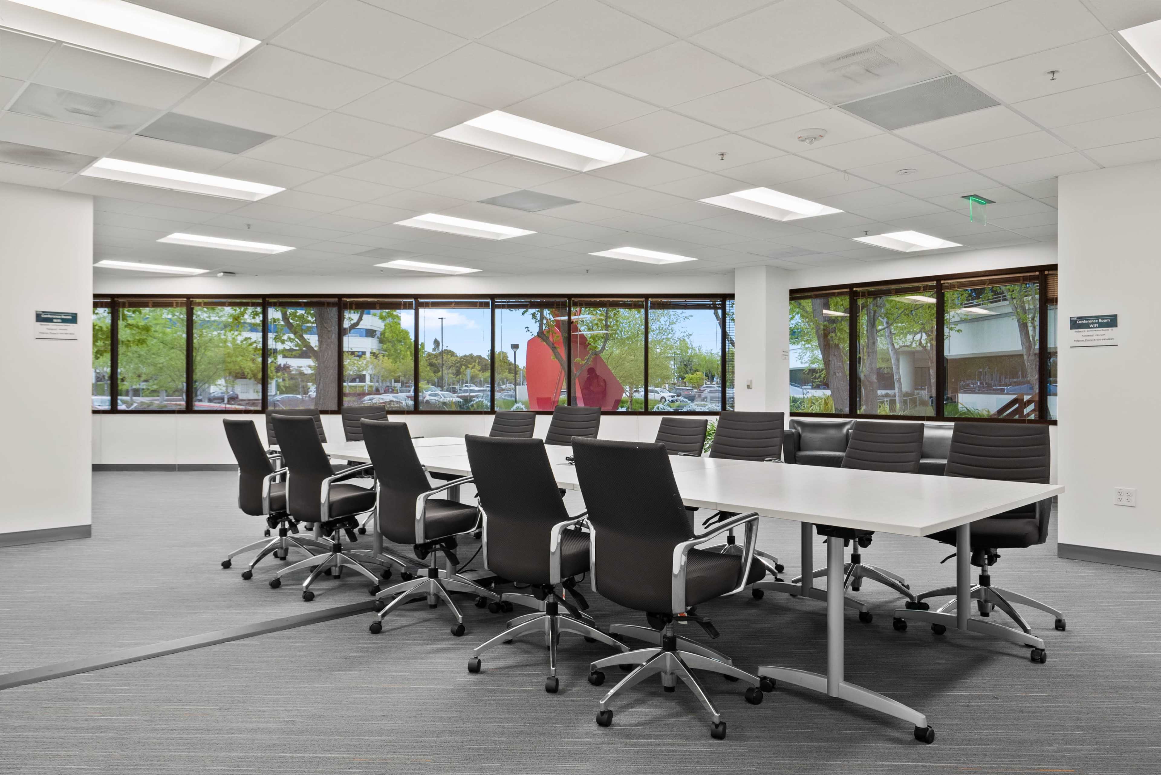 A modern conference room features a long table surrounded by ergonomic chairs, with large windows offering views of green trees and parked cars outside.