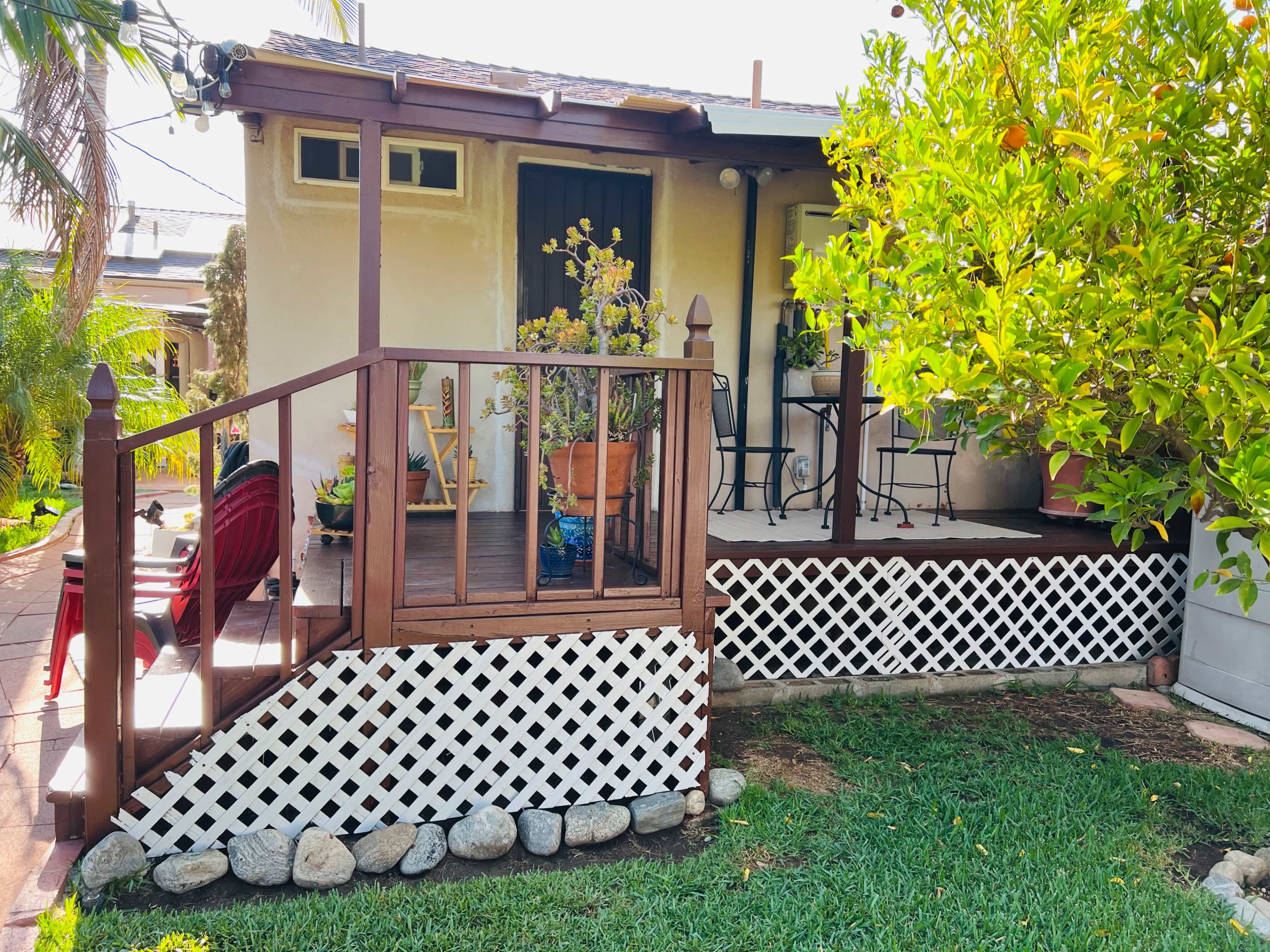 A small house features a wooden deck with a railing, potted plants, and chairs beside a garden with greenery.