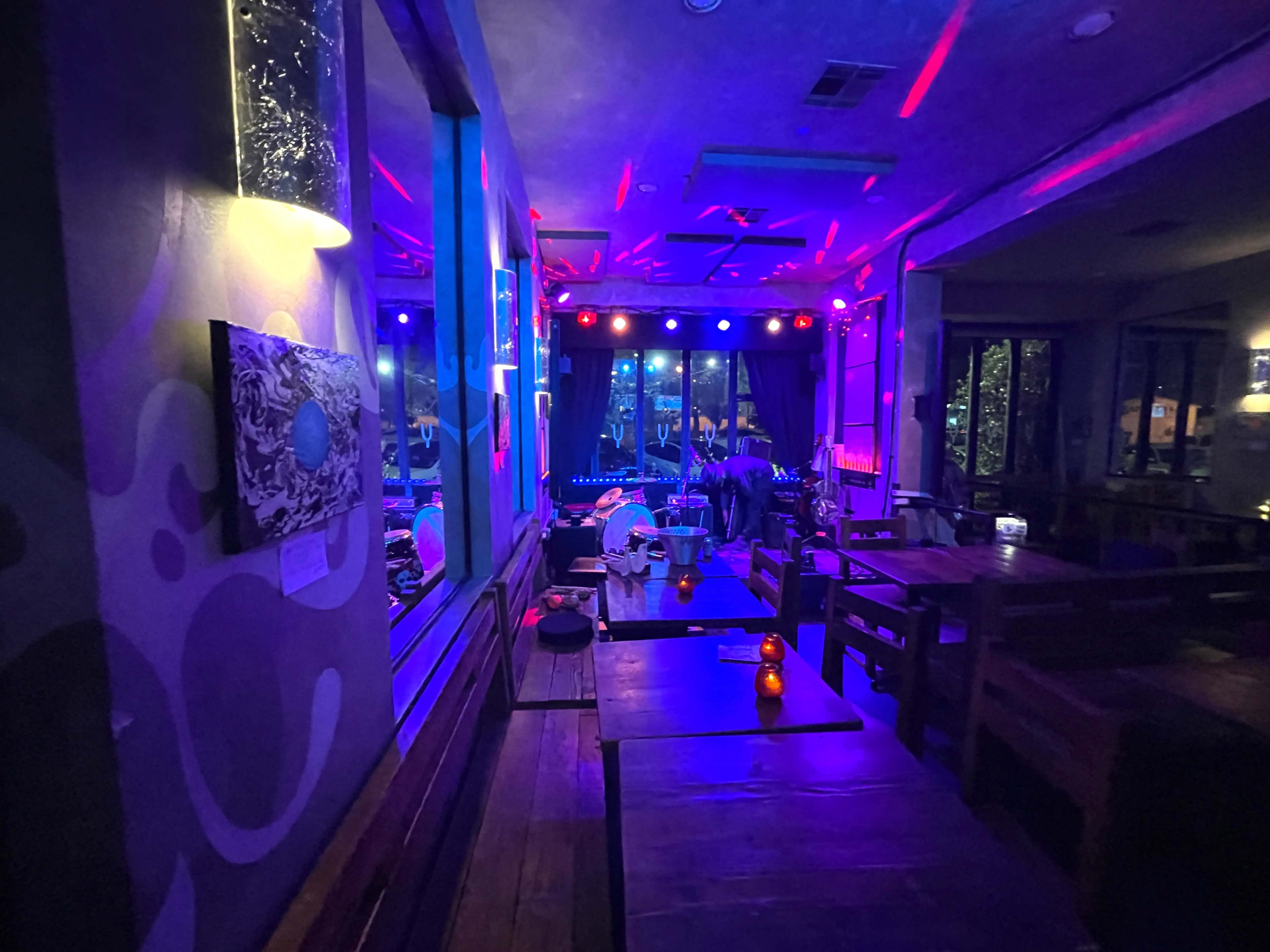 The image shows a dimly lit bar or lounge with colorful lights illuminating a performance stage and empty tables.