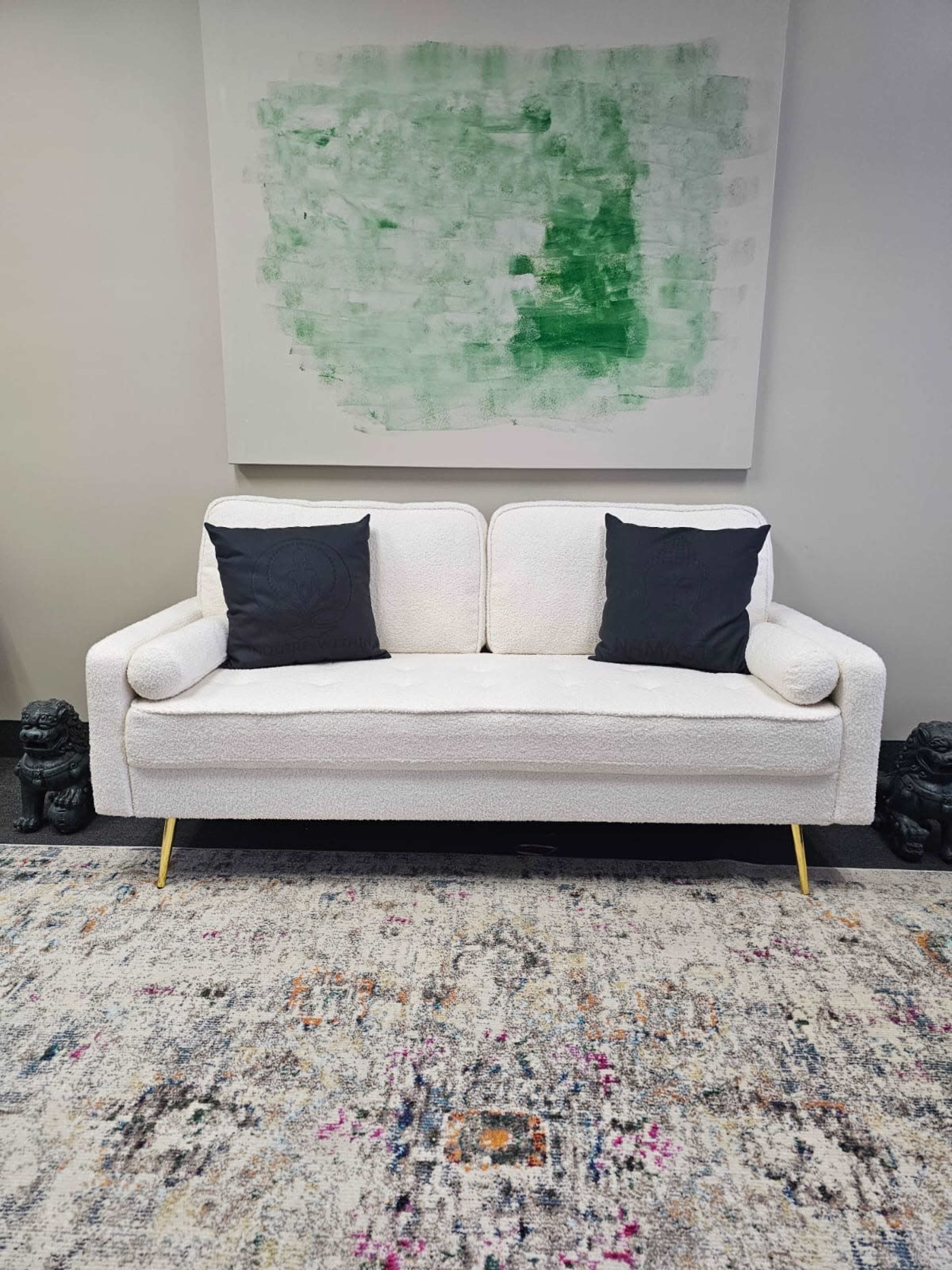 A white sofa with two dark pillows is positioned in front of a green abstract painting on a wall, set against a patterned rug.