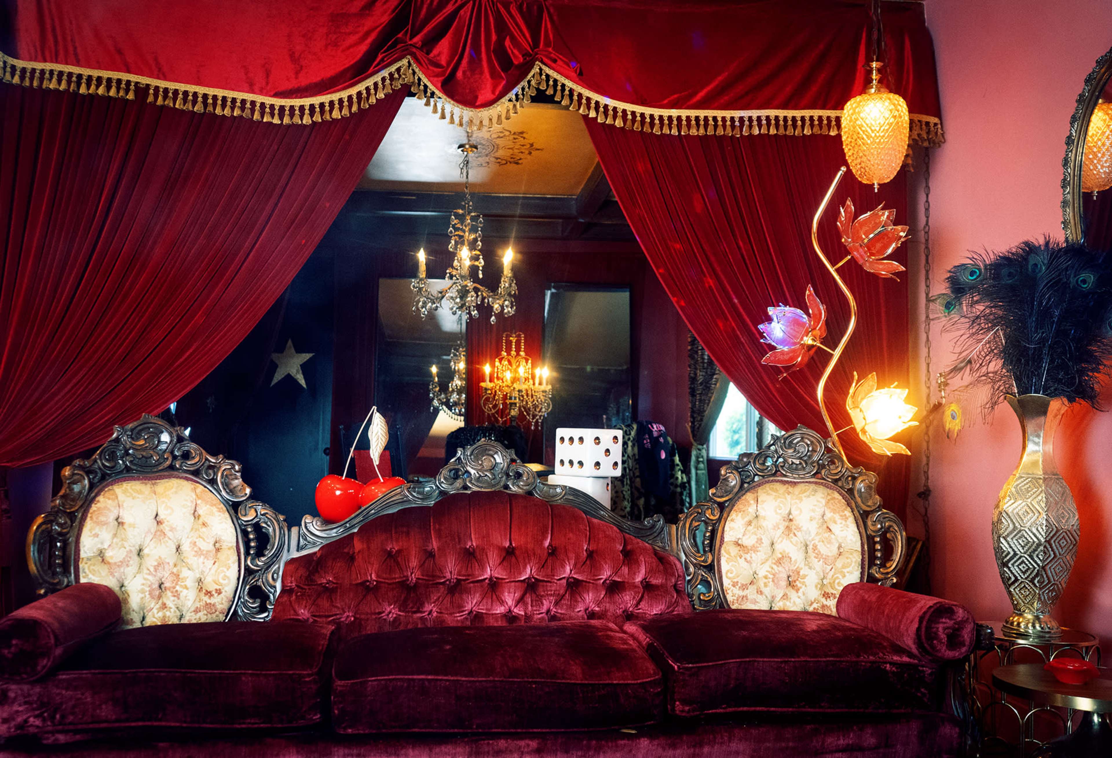 A vintage velvet sofa is positioned against a deep red curtain backdrop, adorned with ornate decorative elements and a chandelier overhead.