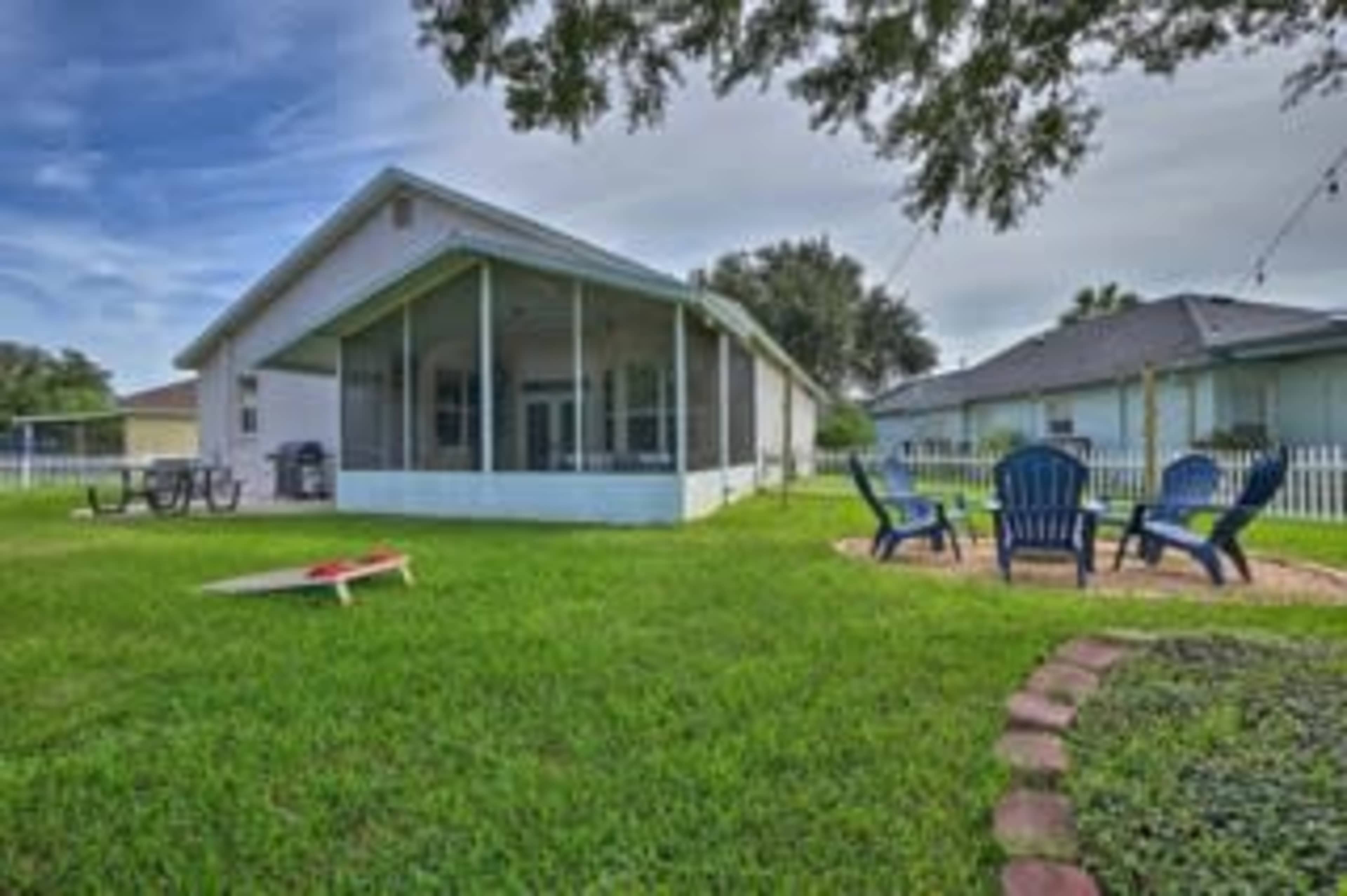 A single-story house with a screened porch sits in a grassy yard, surrounded by a white picket fence and featuring several outdoor seating areas.
