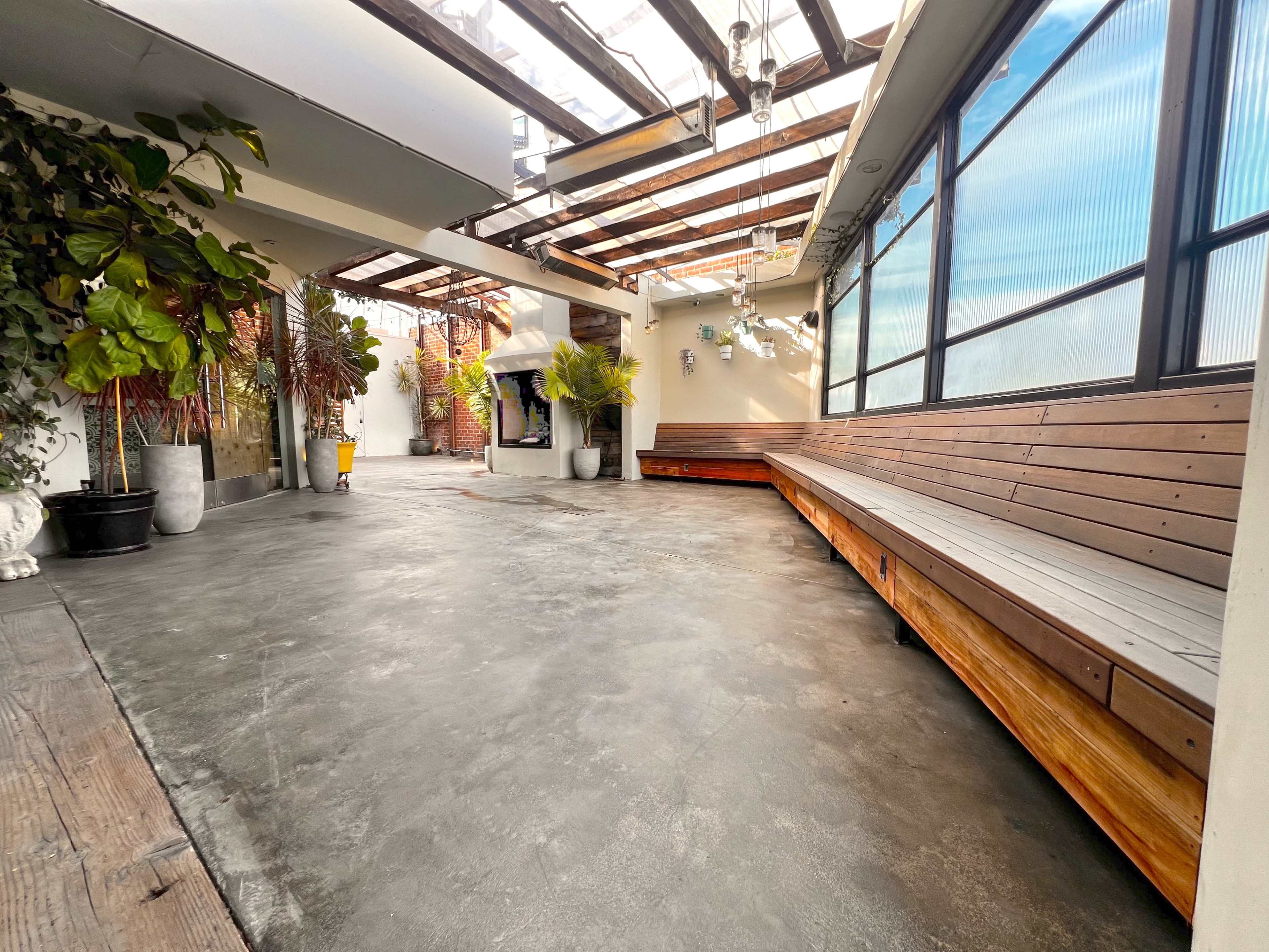 The image shows a spacious, partially covered area with wooden benches, large windows, and potted plants.