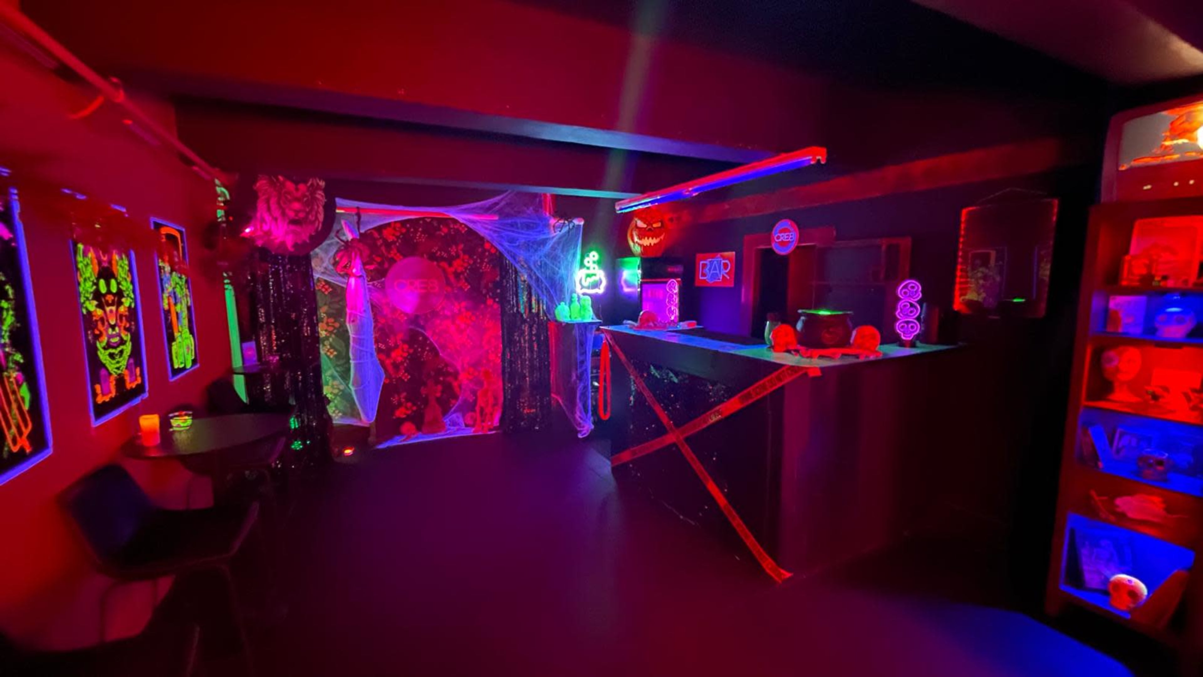 The image shows a dimly lit room decorated for a Halloween-themed event, featuring glowing neon decorations, cobwebs, and pumpkins.