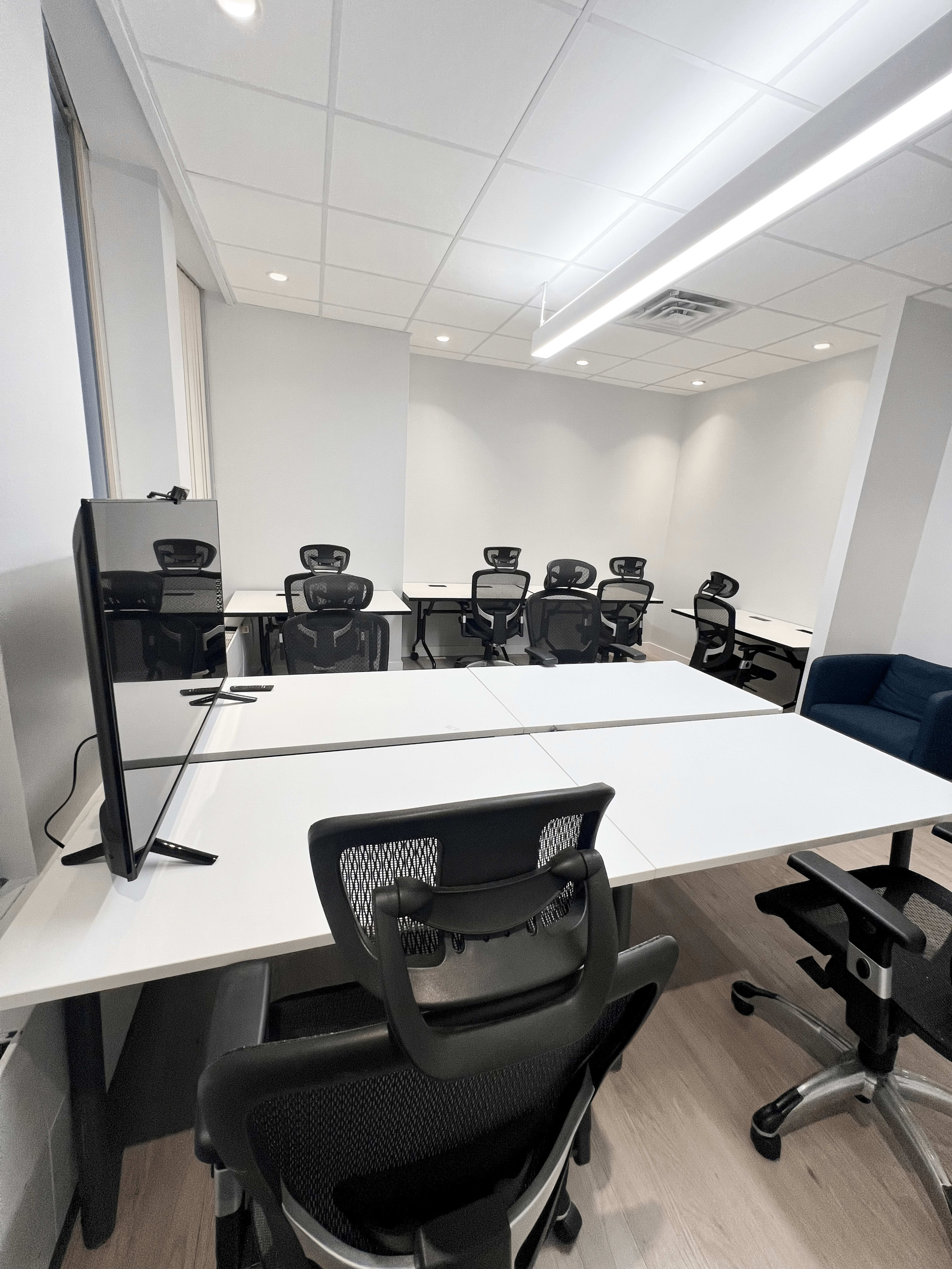 A modern office space features multiple white work tables and ergonomic black chairs arranged in a bright, well-lit room.