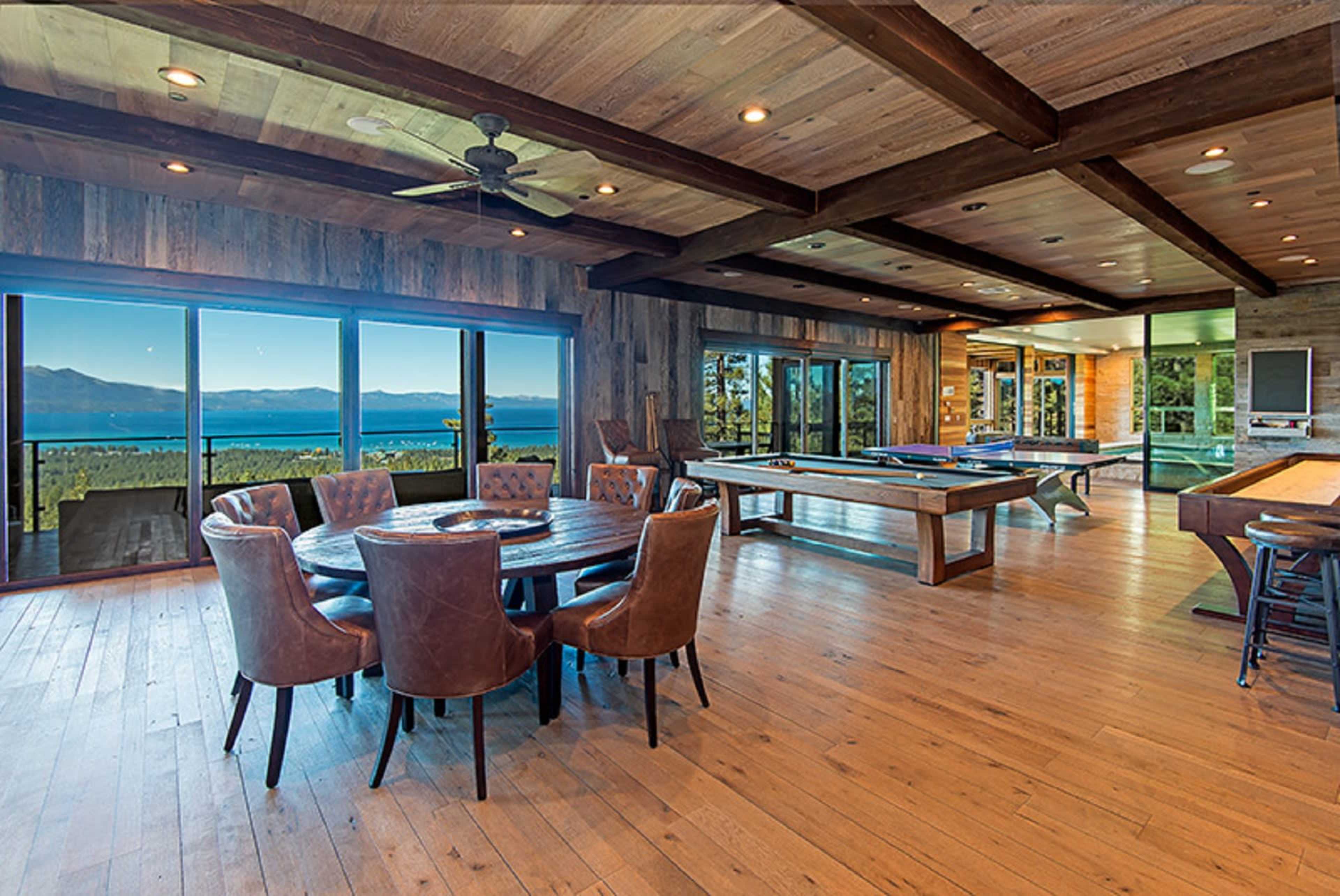 A spacious room featuring wooden floors, large windows with a view of a lake, a dining table with leather chairs, and a game area with a pool table.