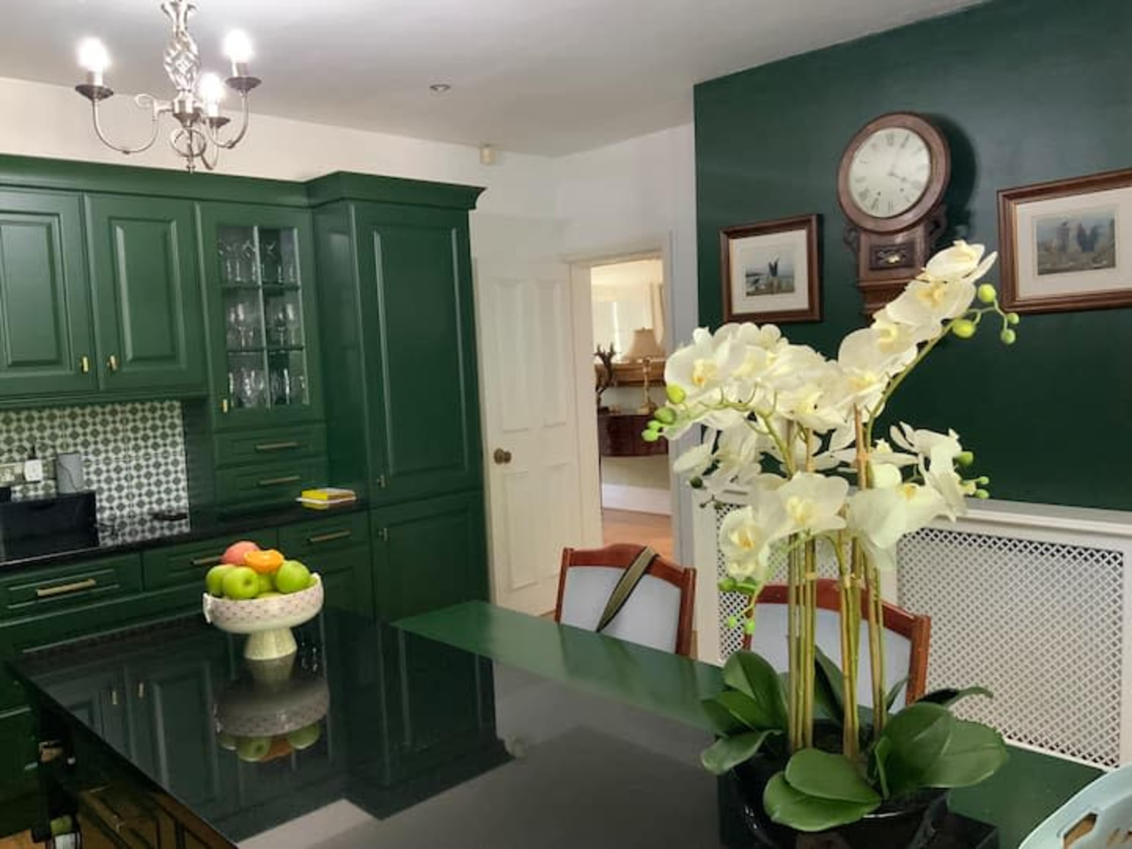 A green-themed kitchen features a central island with a black countertop, a vase of white orchids, and a bowl of fruit.