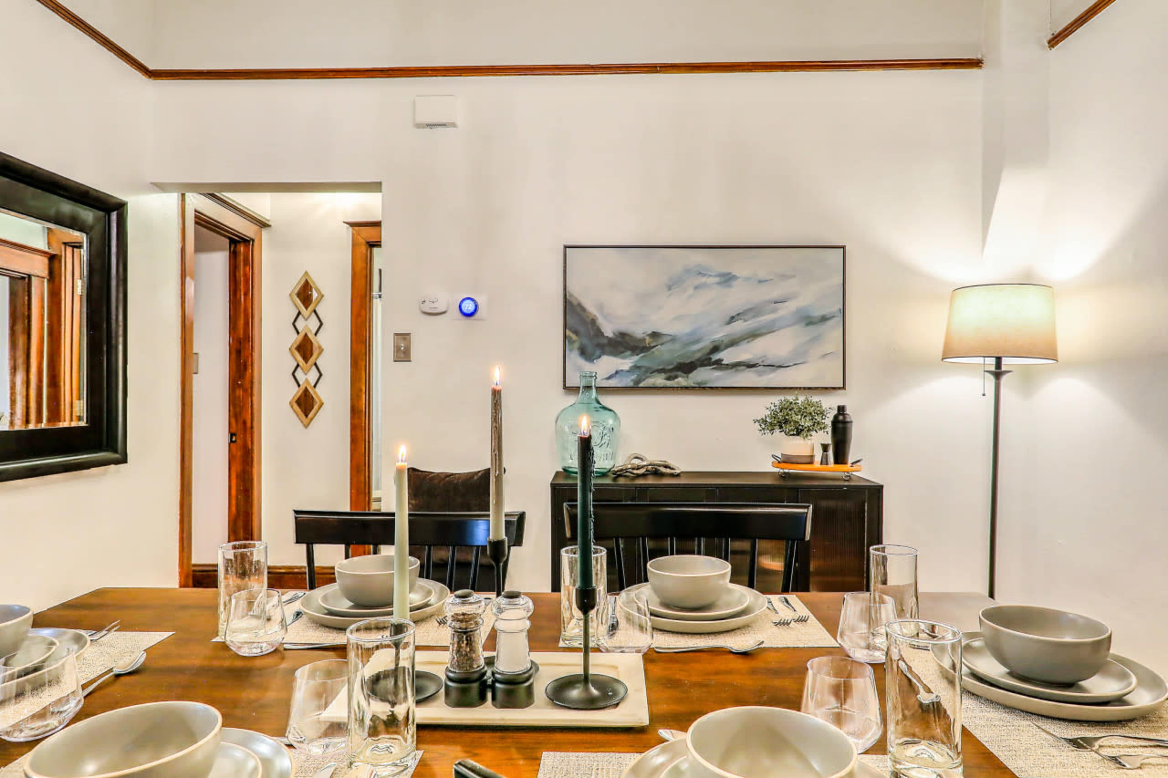 A dining table is set with plates, glasses, and candles, while a painting and a lamp illuminate the room.