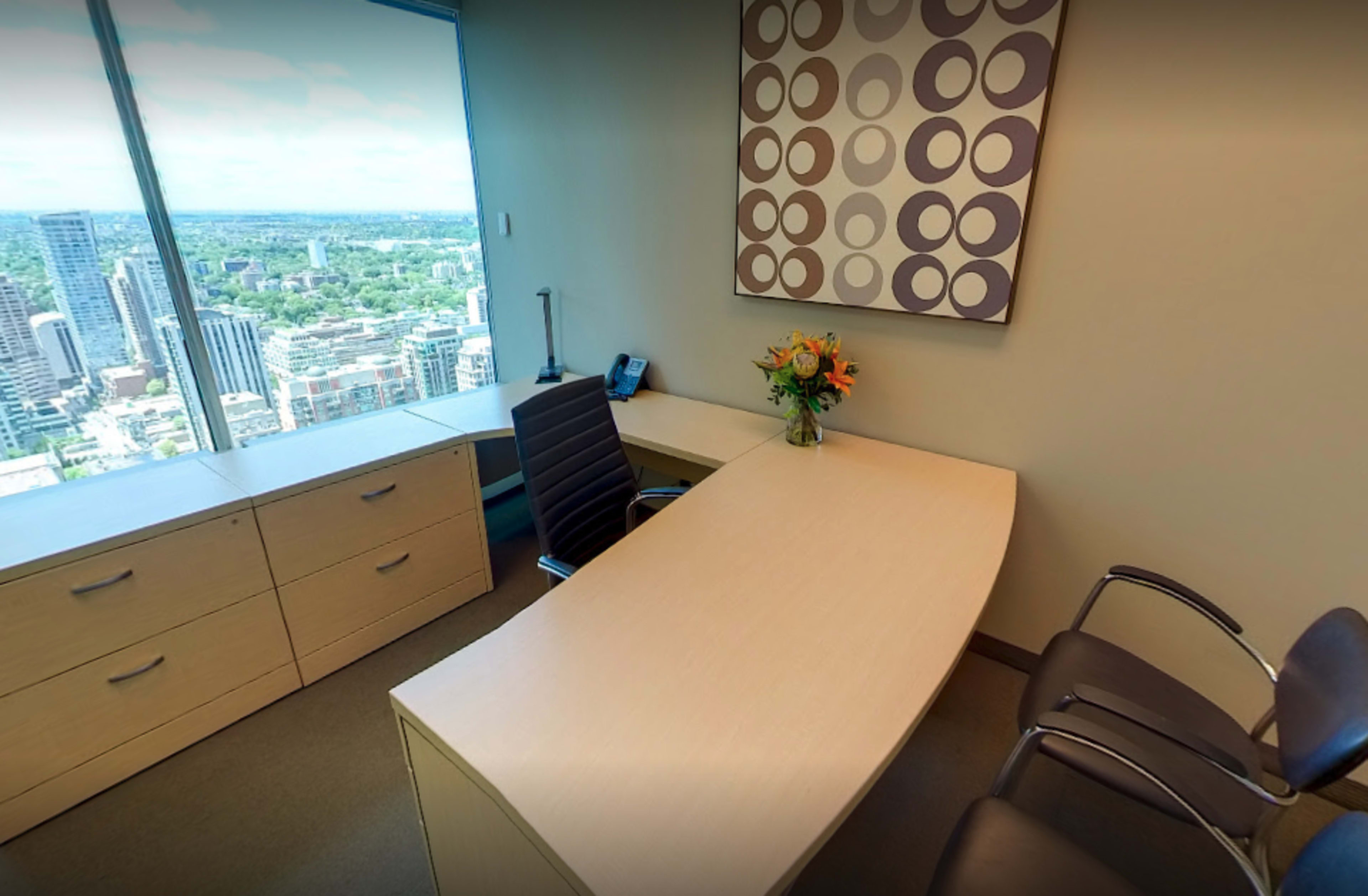 A modern office with a desk, two chairs, a vase of flowers, and a large window overlooking a city skyline.