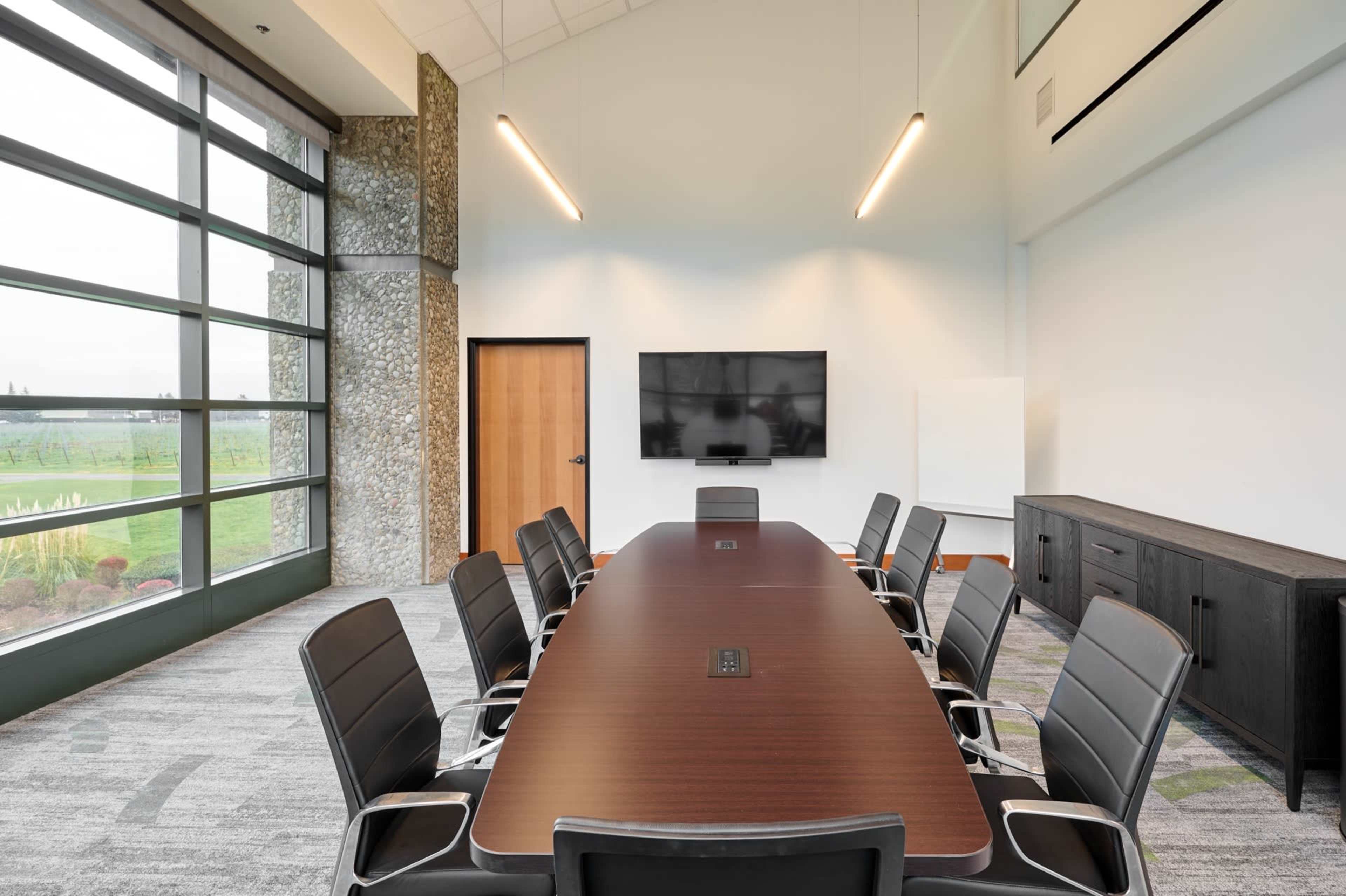 A modern conference room features a long wooden table surrounded by black chairs, a large screen on the wall, and large windows allowing natural light to illuminate the space.