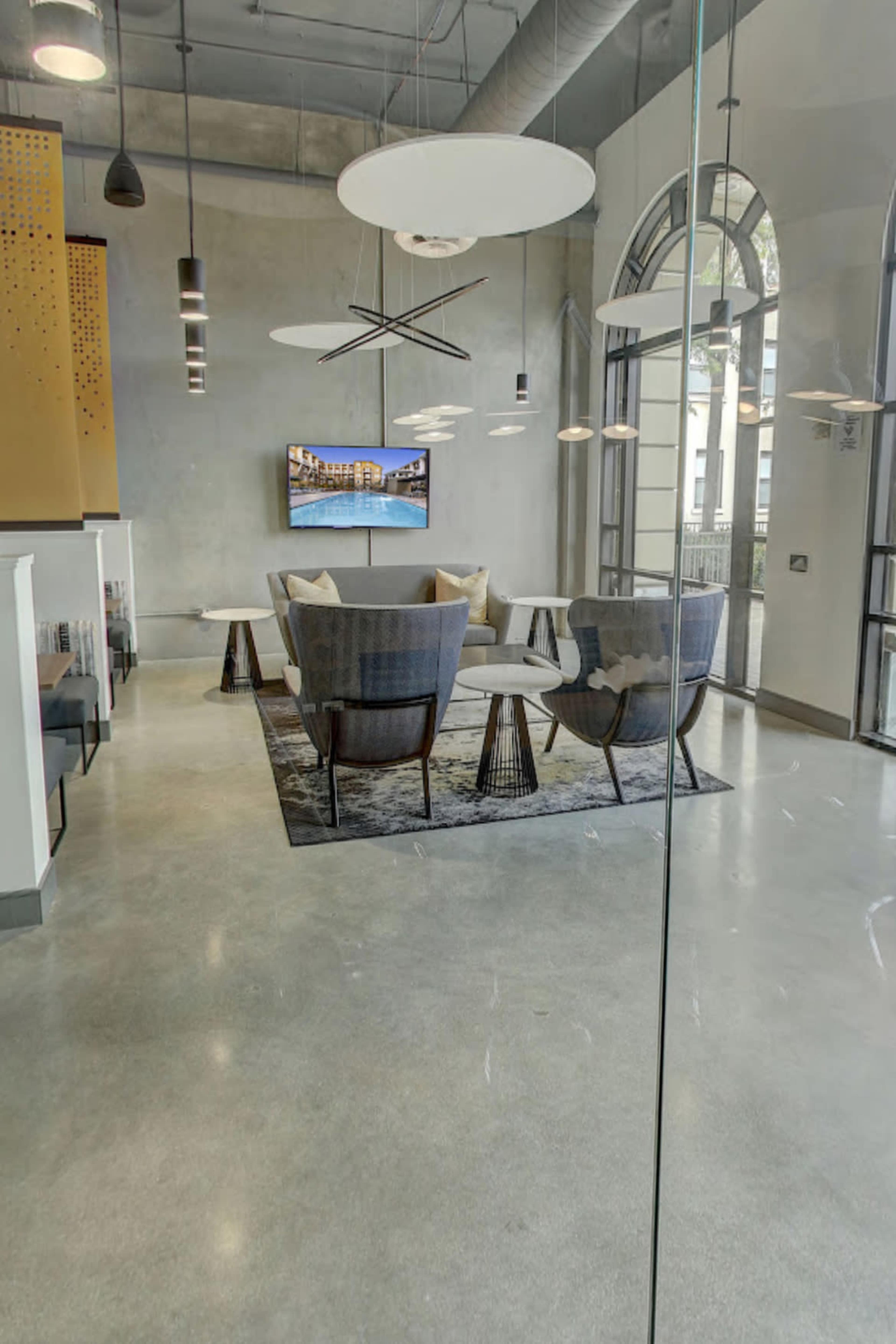 A modern lobby area featuring a seating arrangement with gray chairs and a round table, illuminated by pendant lights and a large television on the wall.