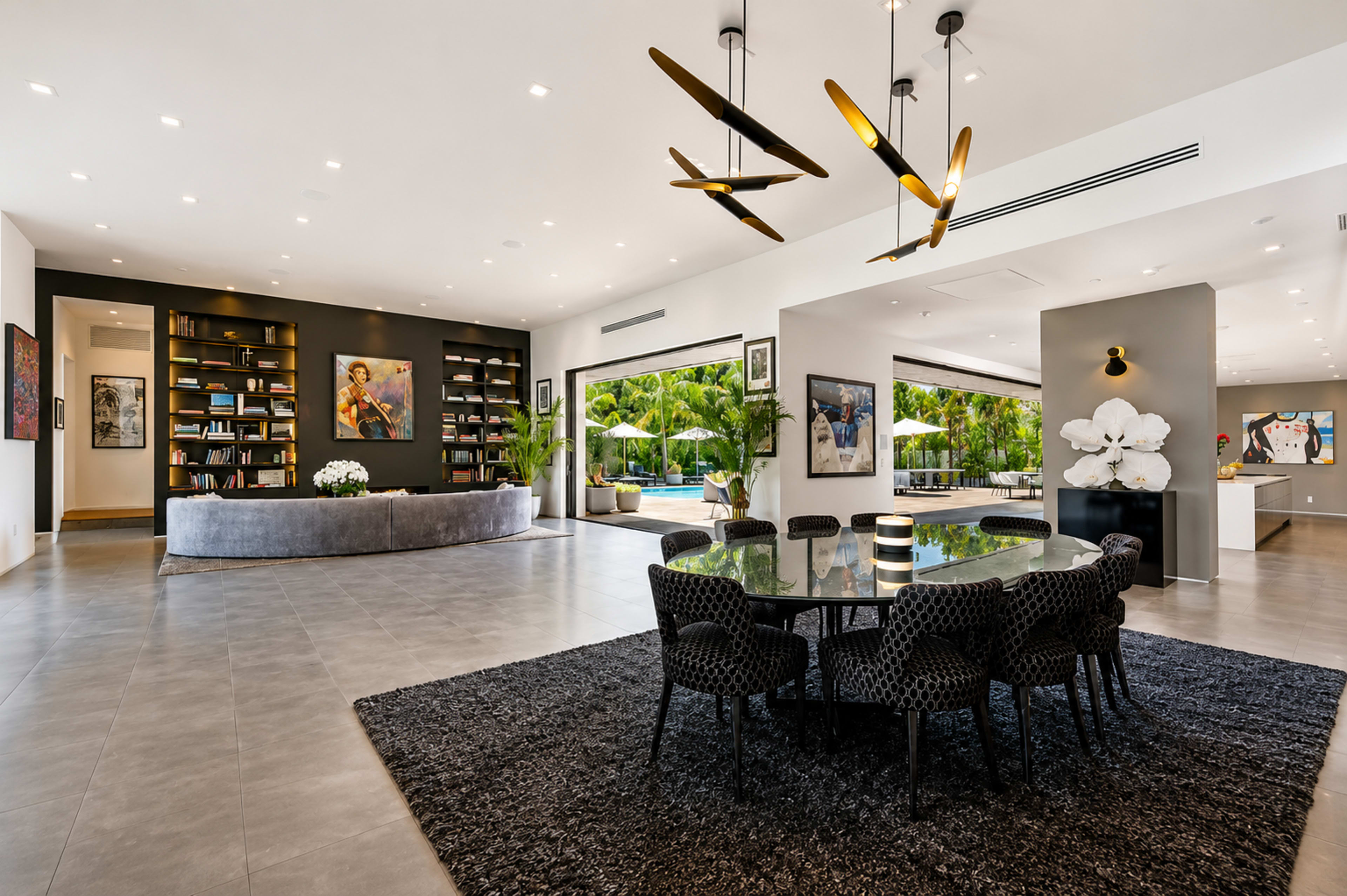 The image shows a modern open-concept living space featuring a dining area with a large round table surrounded by black chairs, a curved bookshelf, and floor-to-ceiling windows overlooking a patio with greenery.