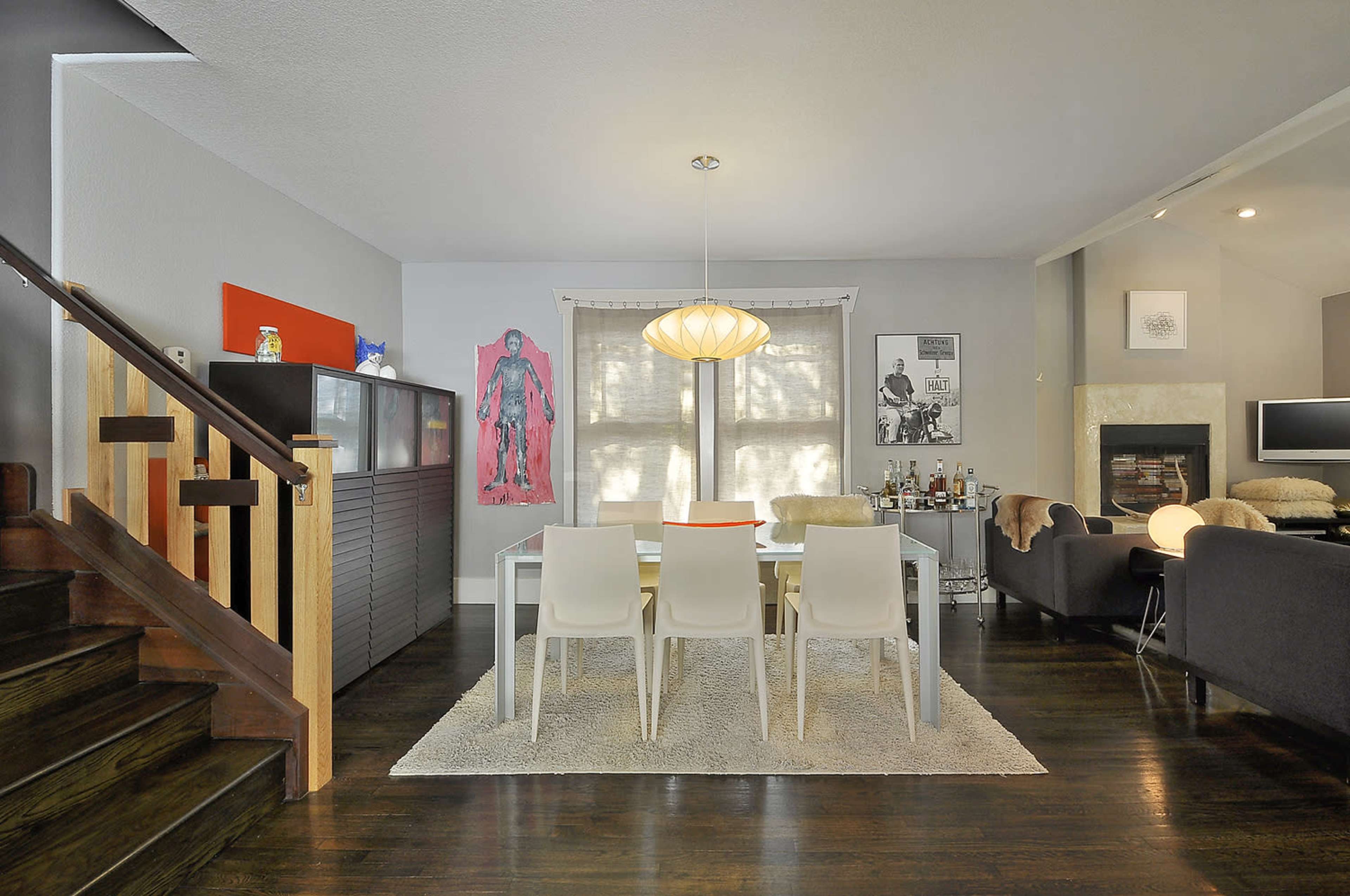 A modern dining area featuring a white table surrounded by chairs, with a staircase on one side and a cozy seating area opposite, illuminated by a pendant light.