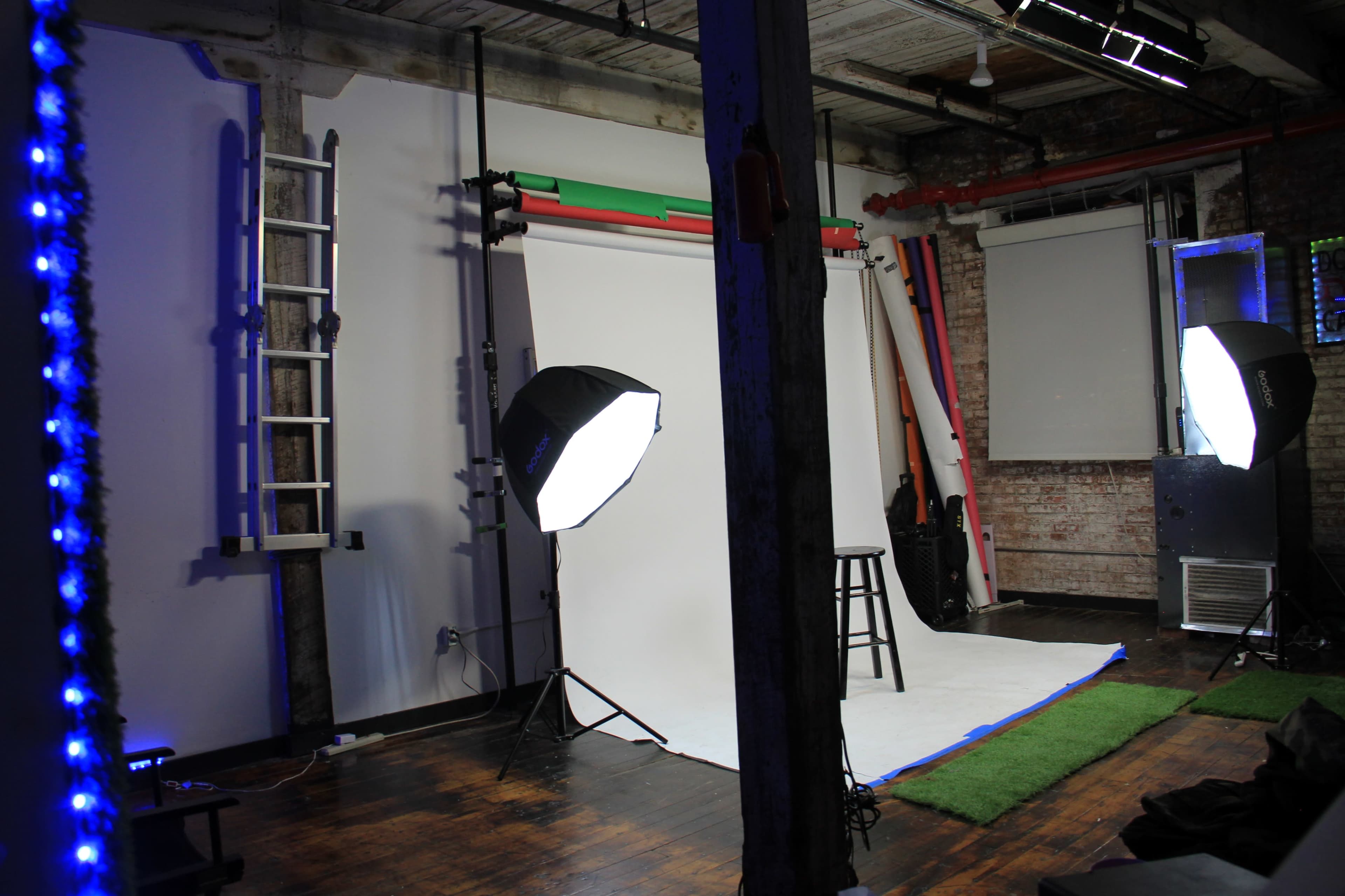 A photography studio features a white backdrop with lighting equipment, a stool, and a ladder against the wall.
