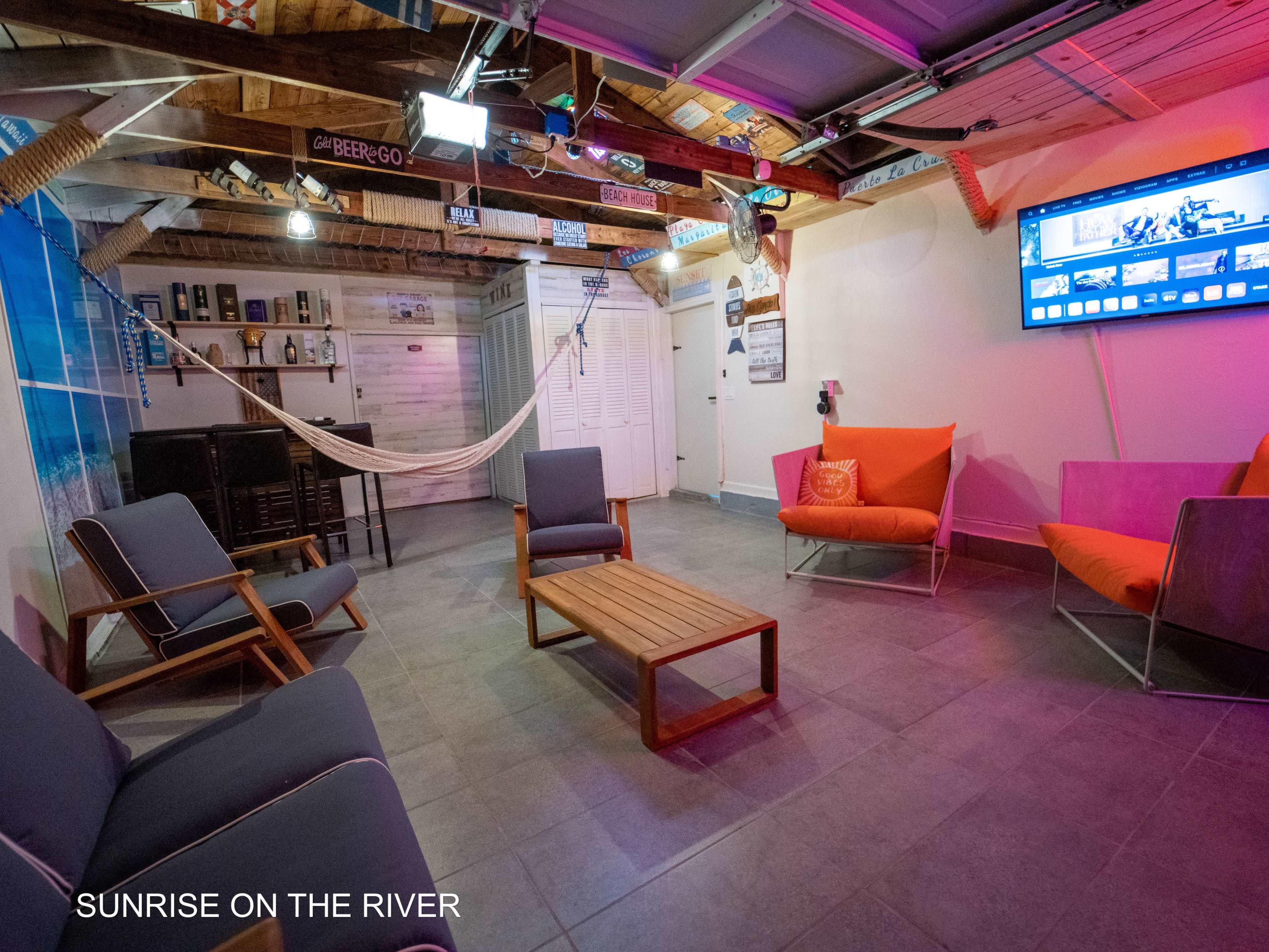 A cozy indoor space features lounge seating, a coffee table, a hammock, and a wall-mounted television under a wooden ceiling with exposed beams.