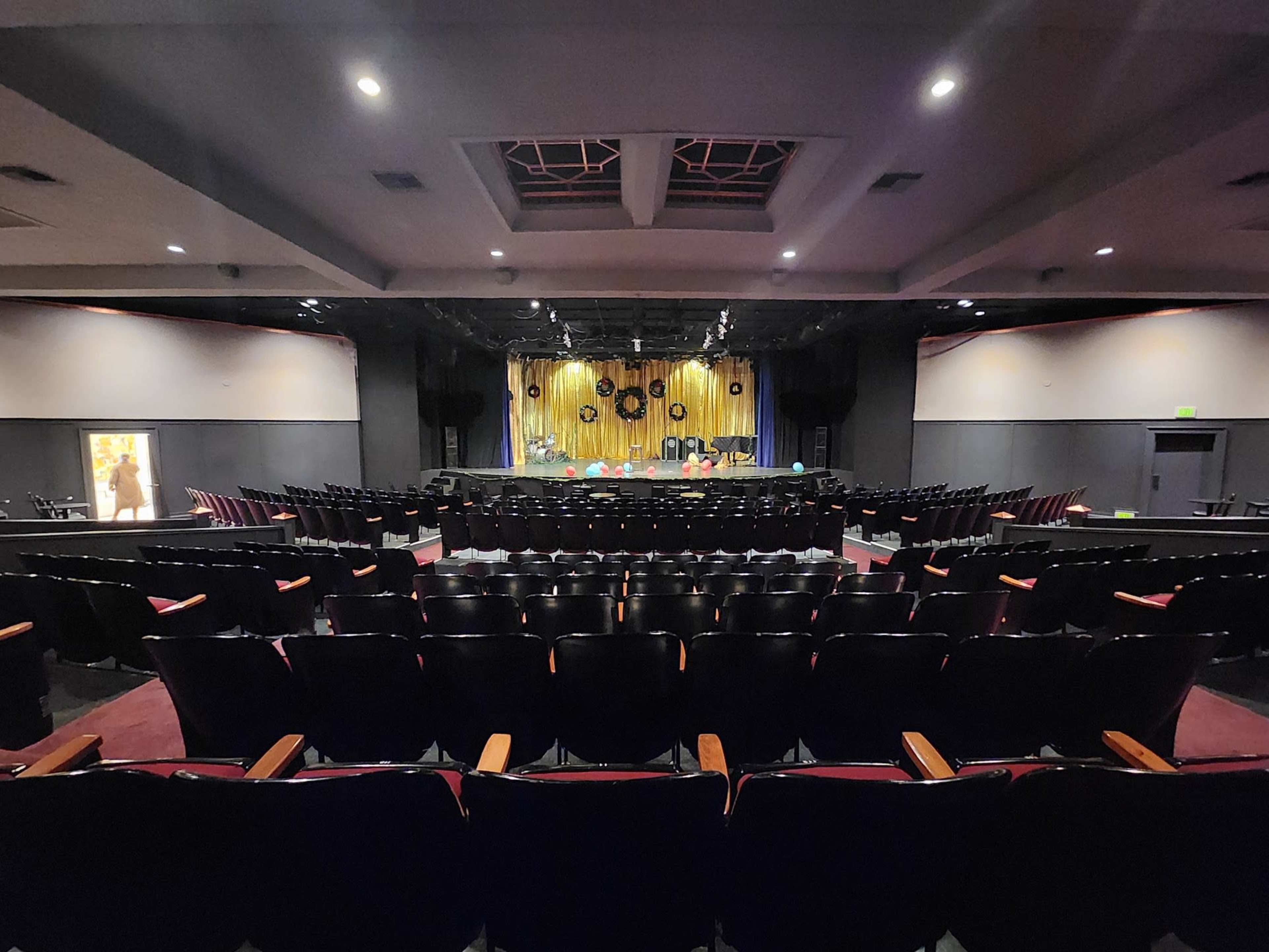 A darkened theater with rows of empty black seats facing a stage adorned with yellow curtains and decorative elements.
