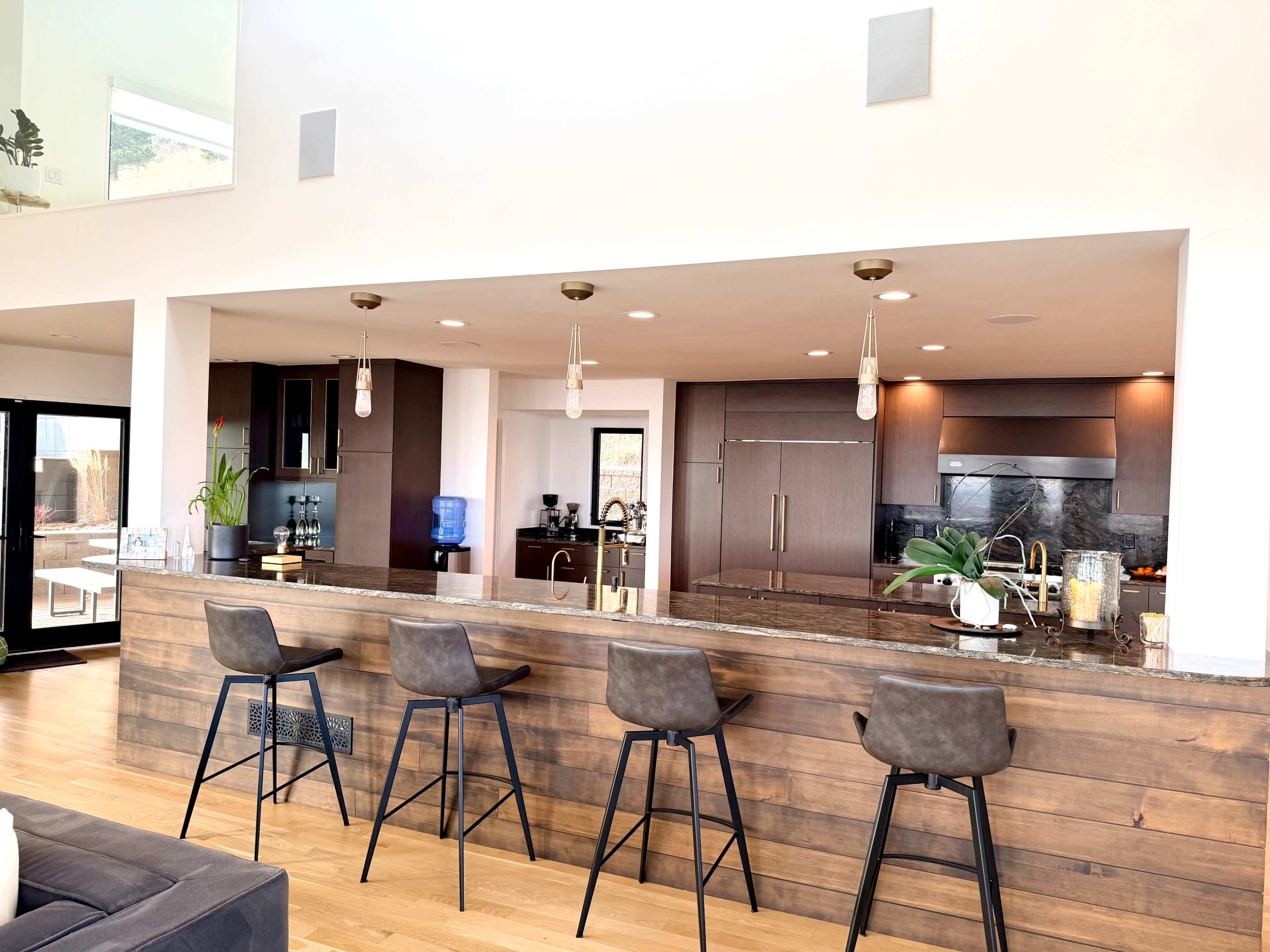 The image shows a modern kitchen with a wooden bar area featuring several stools, sleek countertops, and high-end appliances.