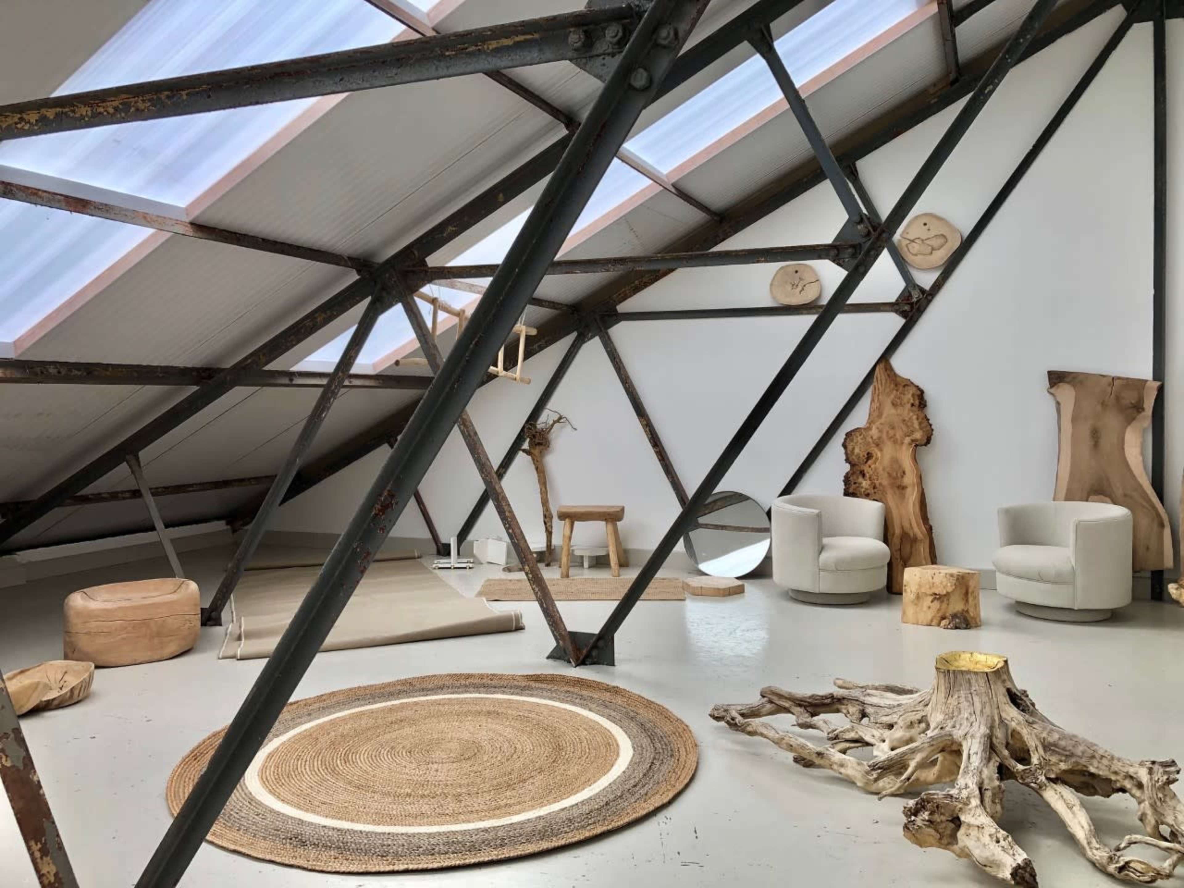 A minimalist attic space with large windows, featuring wooden decor, two white chairs, a round rug, and exposed structural beams.