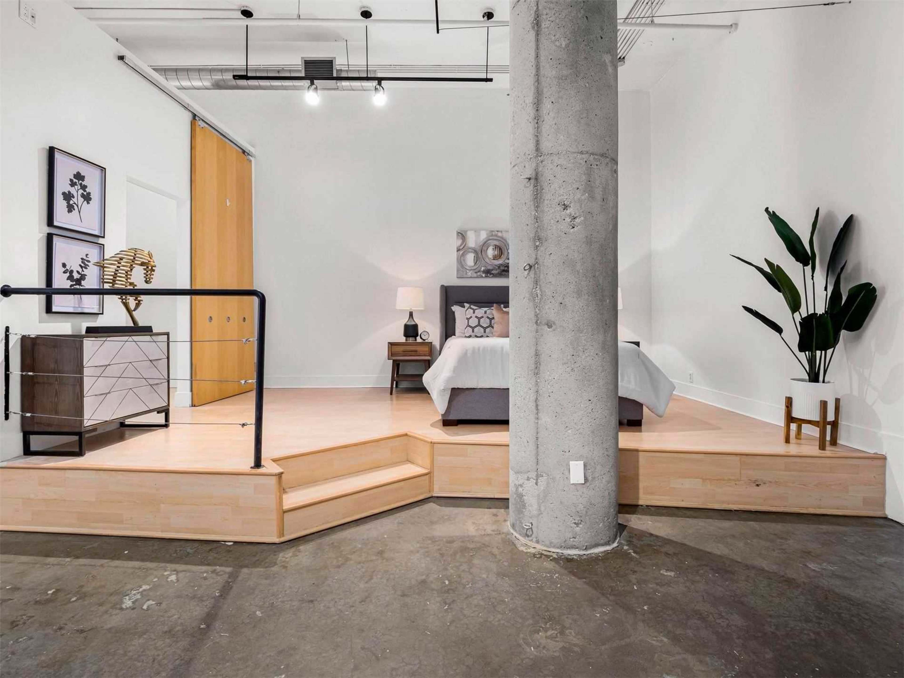 A modern bedroom features a raised platform bed with decorative elements and a large concrete pillar, set against a minimalist backdrop.
