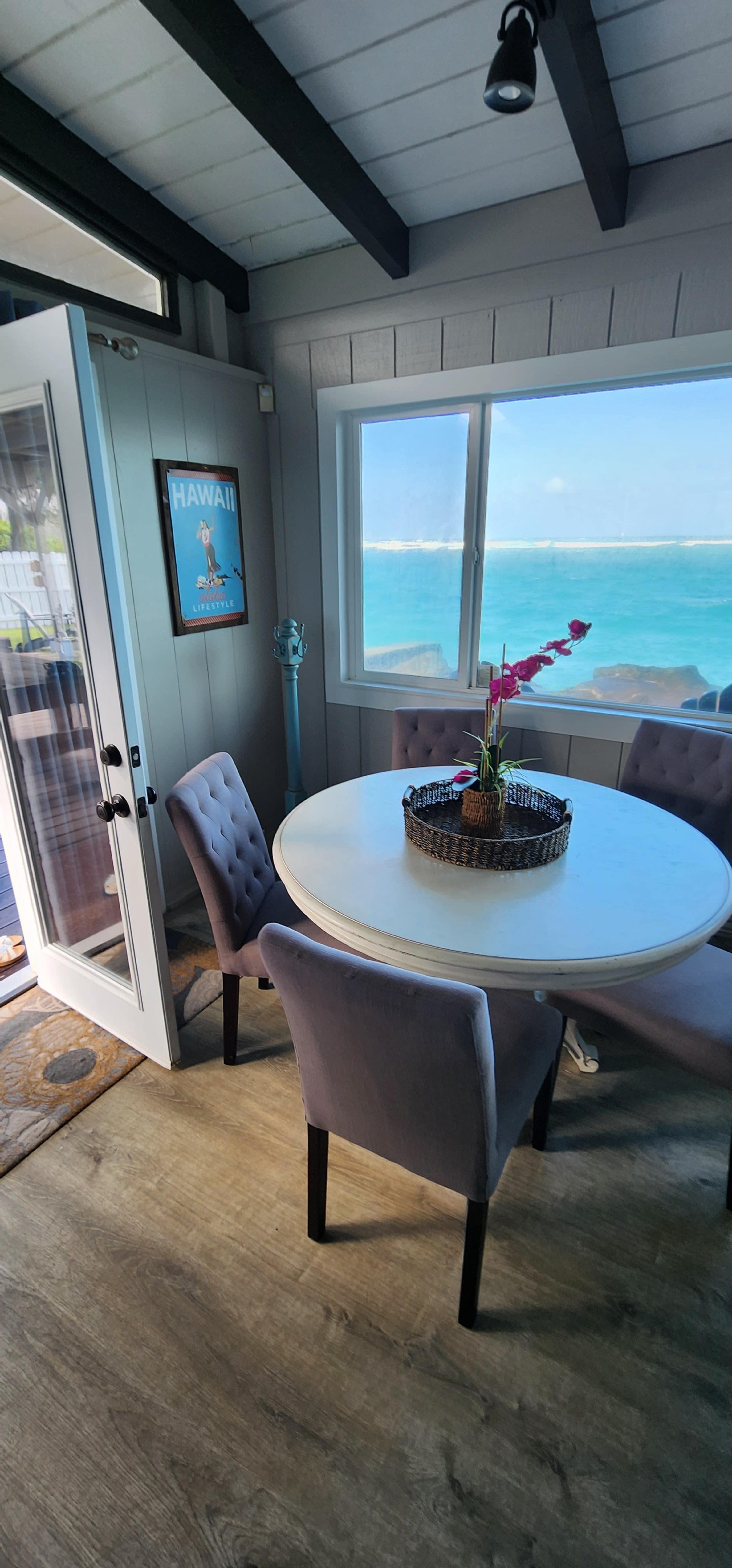 The image shows a dining area with a round table and four upholstered chairs, adjacent to a large window offering a view of the ocean.