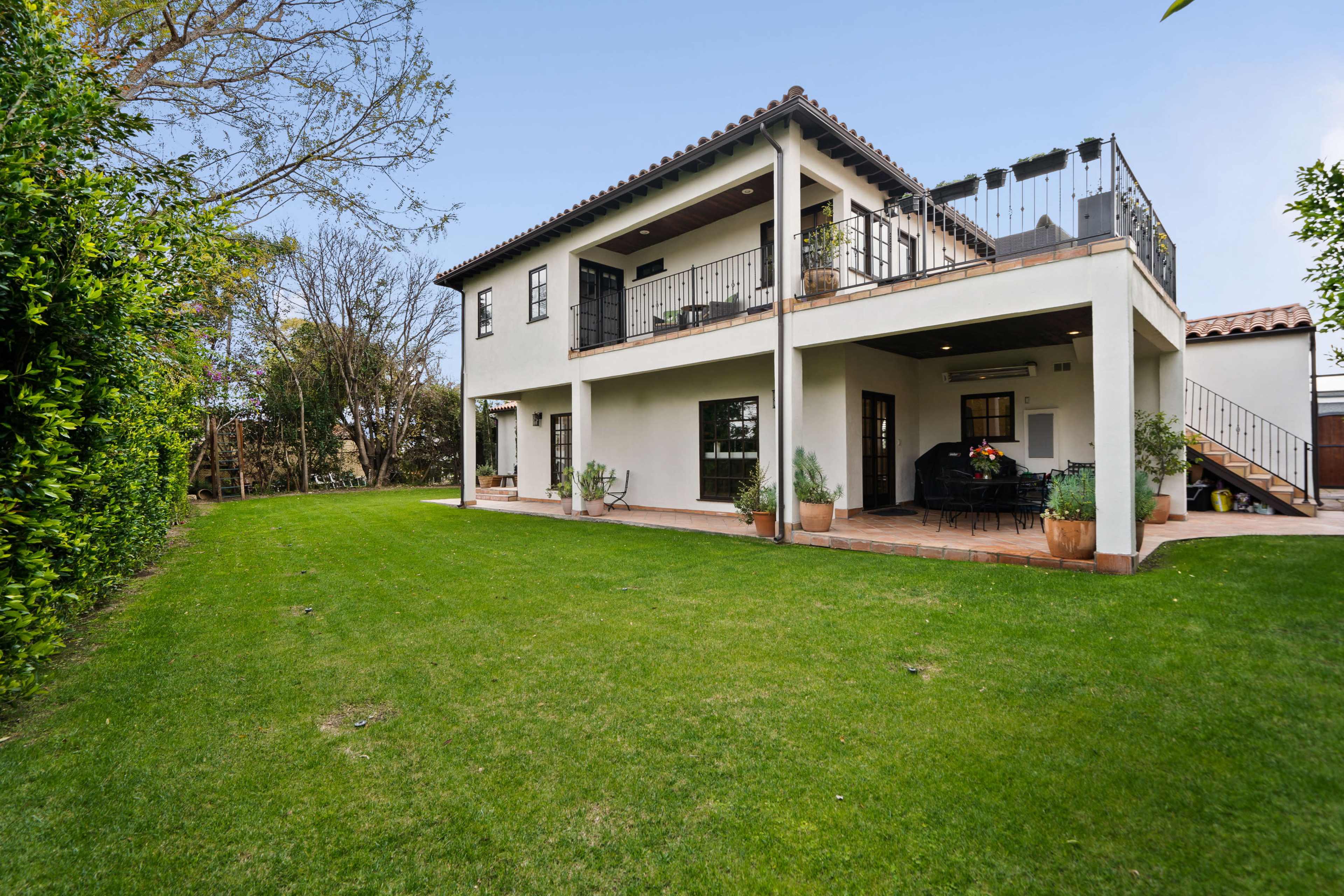 A two-story house with a tiled roof has a balcony and opens to a grassy backyard surrounded by shrubs and trees.