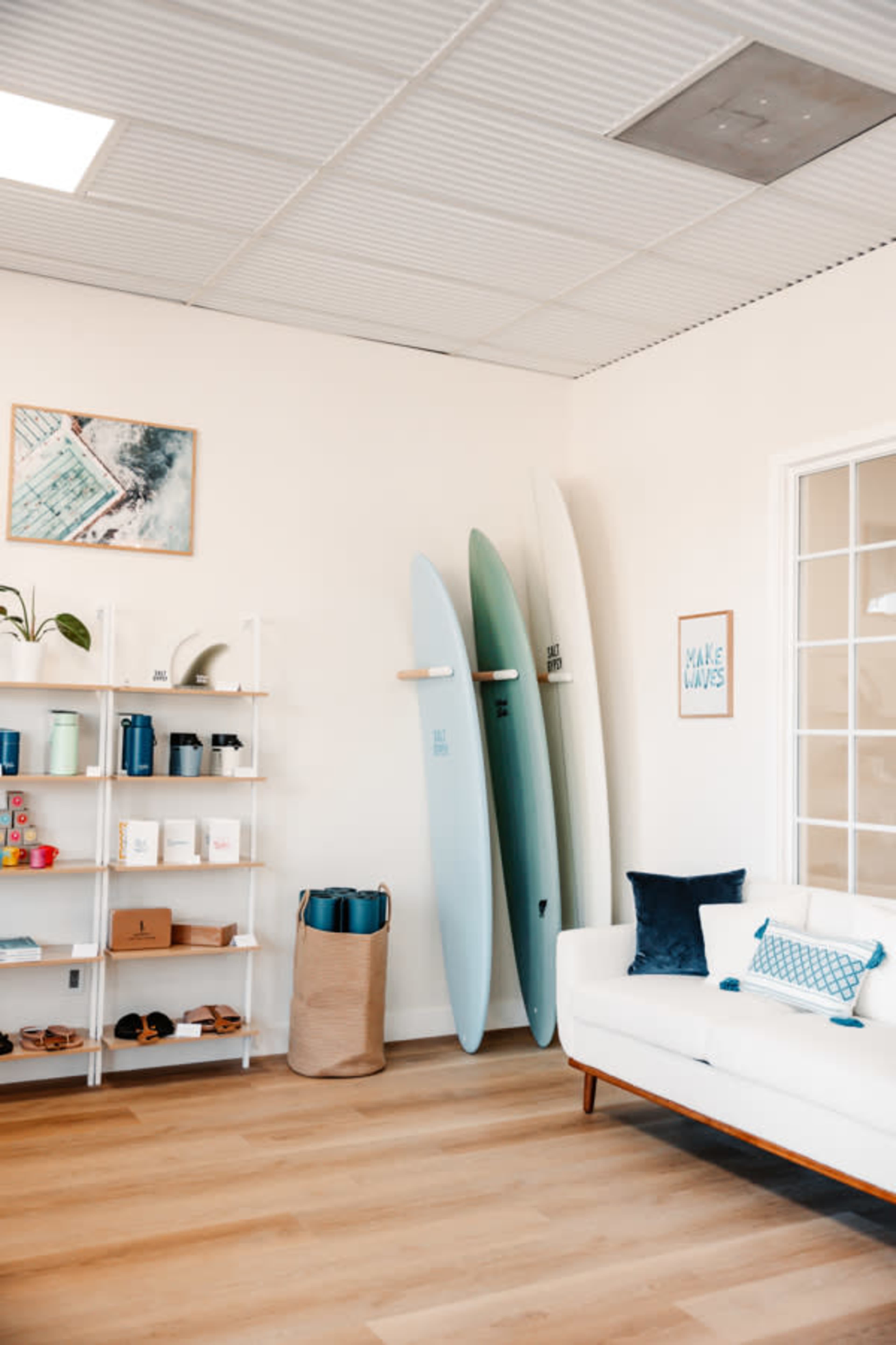 The image shows a bright, modern room featuring three surfboards mounted on the wall, a white couch, a shelf with various items, and a decorative artwork.