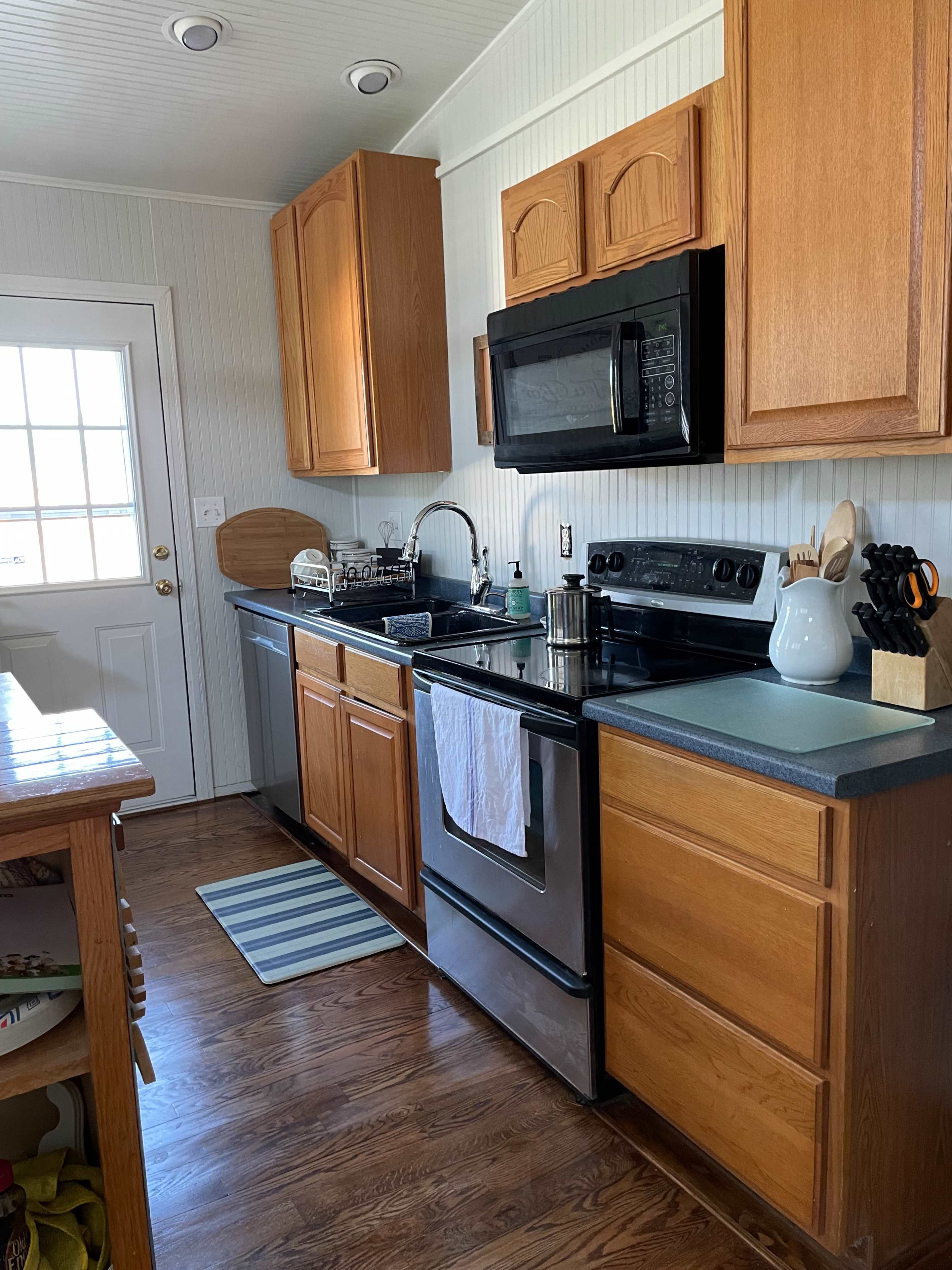 A kitchen with wooden cabinets, a black stove and microwave, a stainless steel sink, and a door leading outside.
