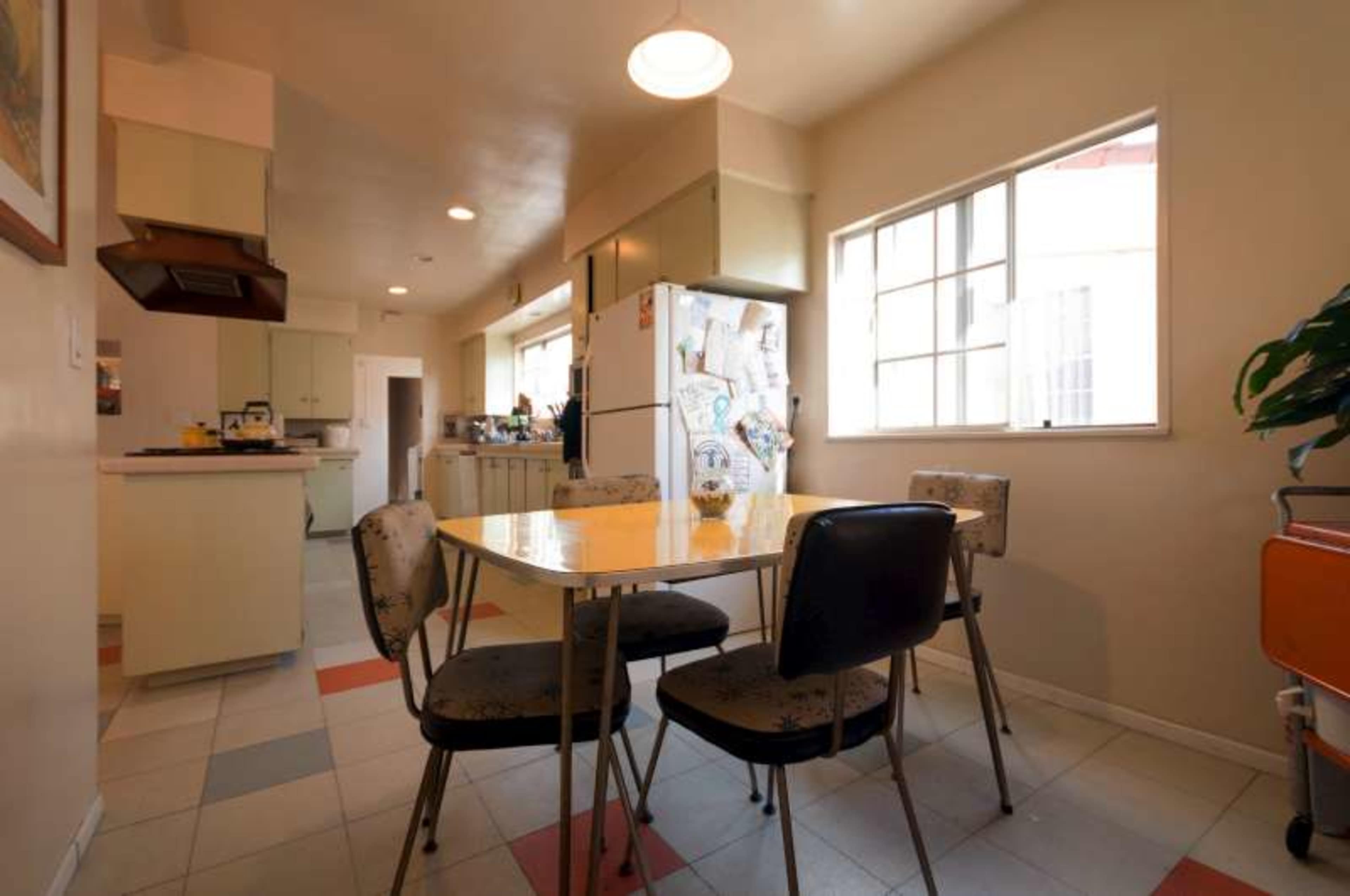 The image shows a kitchen with a small dining table and four chairs, a refrigerator, and windows allowing natural light to enter.