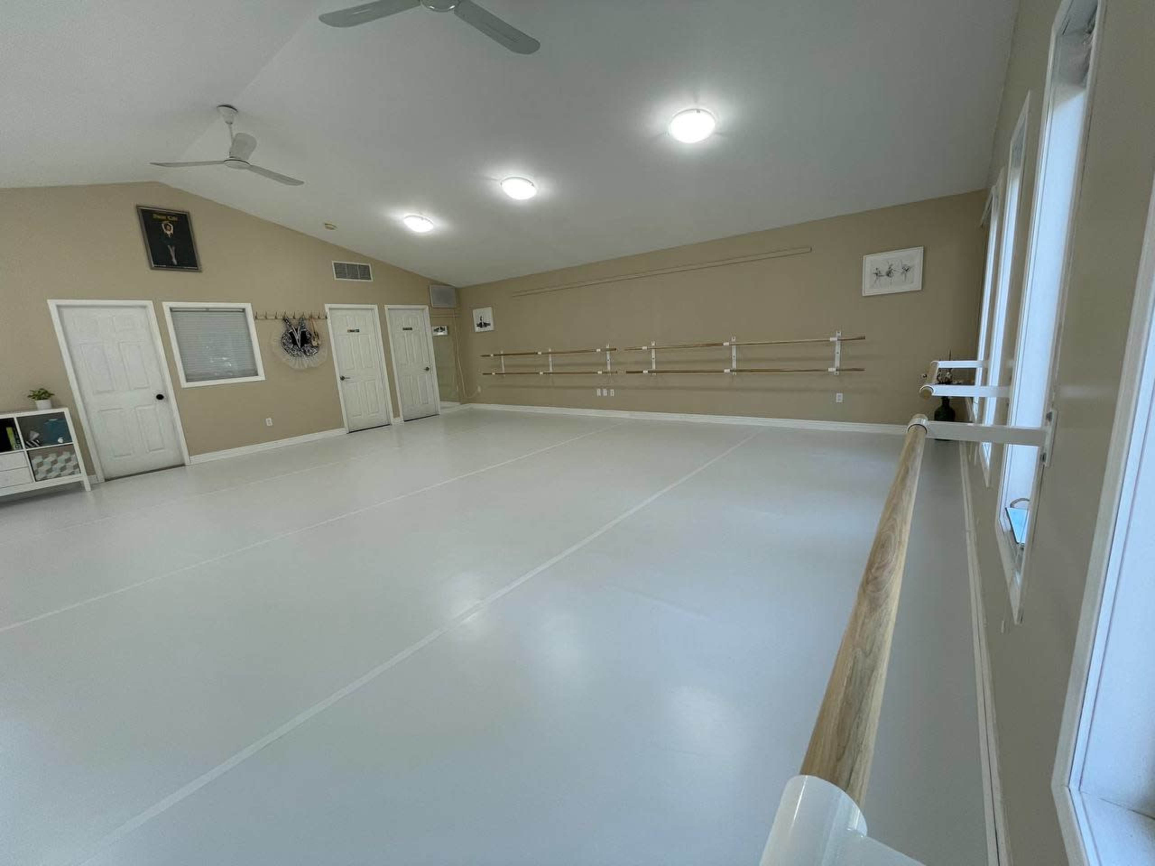 A spacious dance studio with a clean, white floor, ballet bars mounted on the walls, and large windows providing natural light.