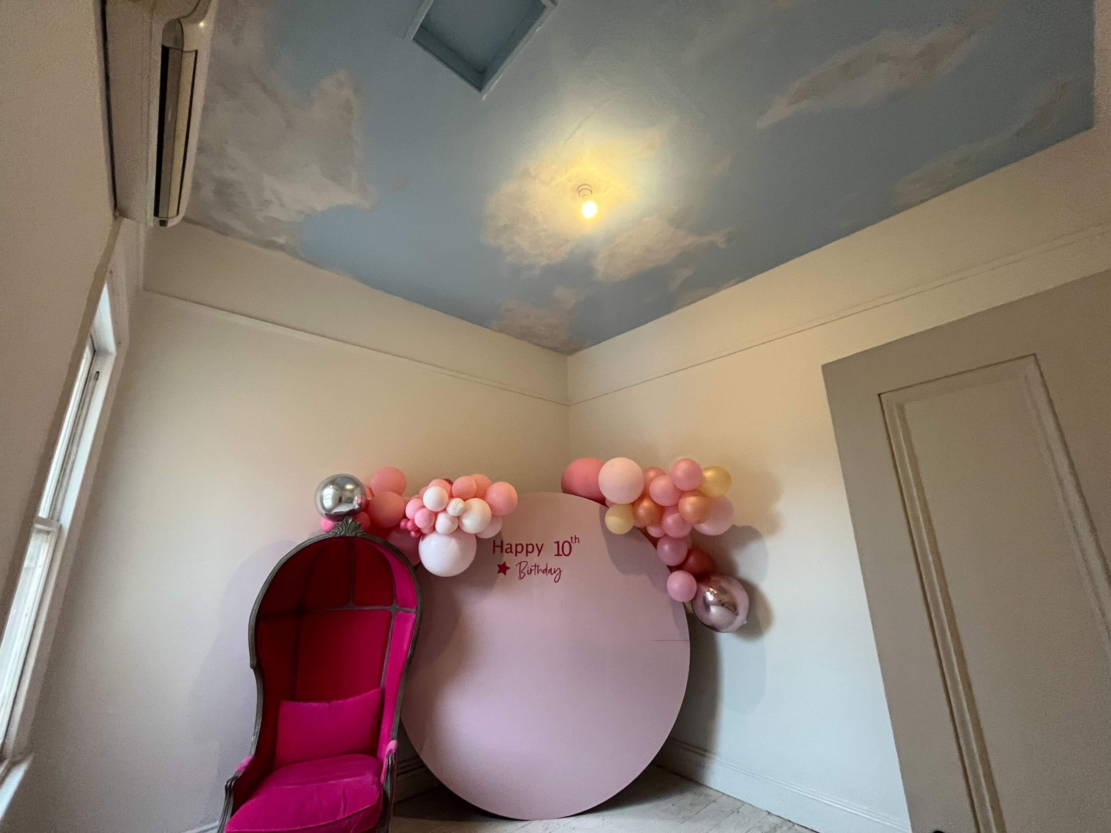 A room with a sky-painted ceiling, a pink chair, and a round backdrop adorned with balloons and a birthday message.