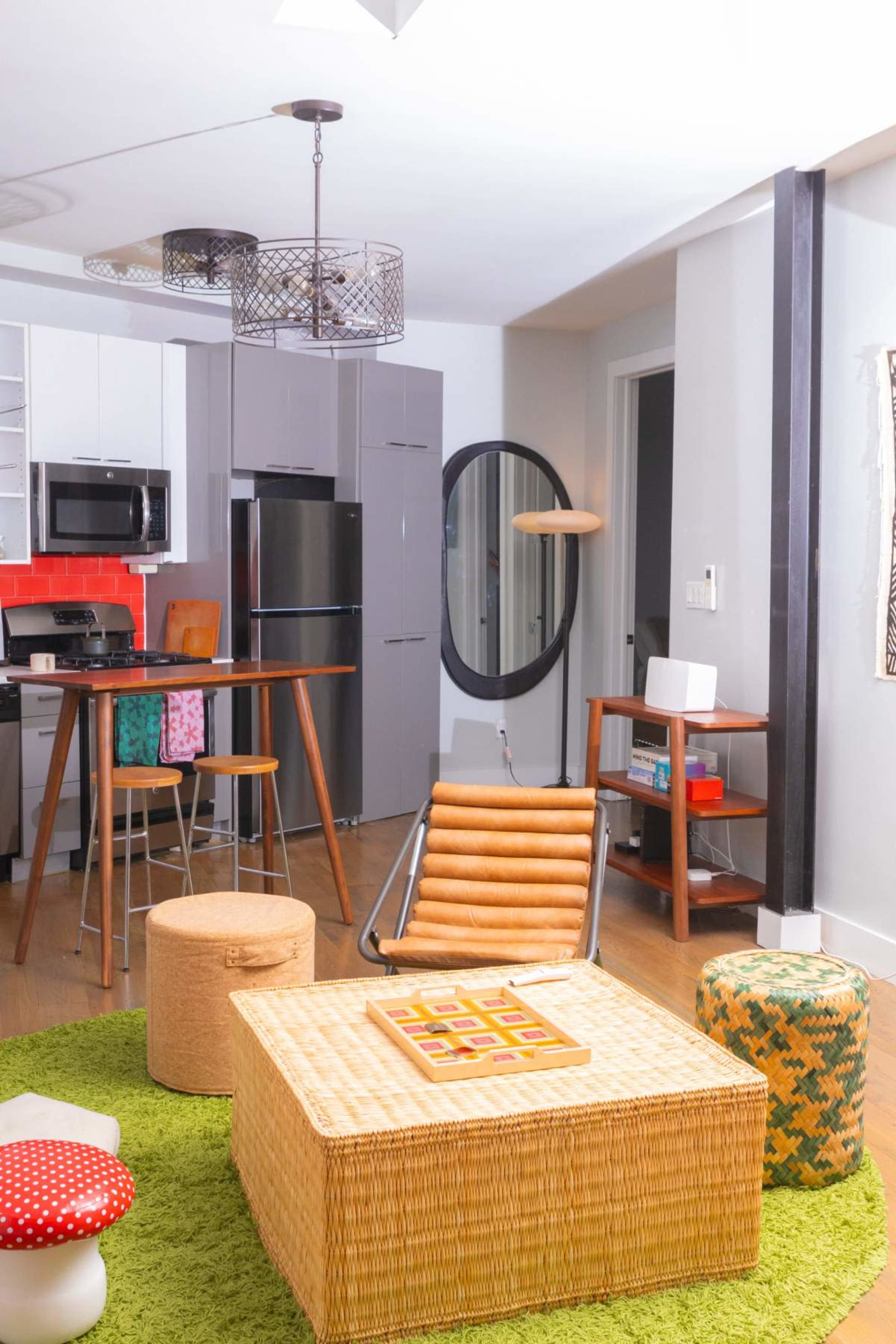 The image shows a modern living space featuring a kitchenette with stainless steel appliances, a circular mirror, and a variety of seating options on a green area rug.