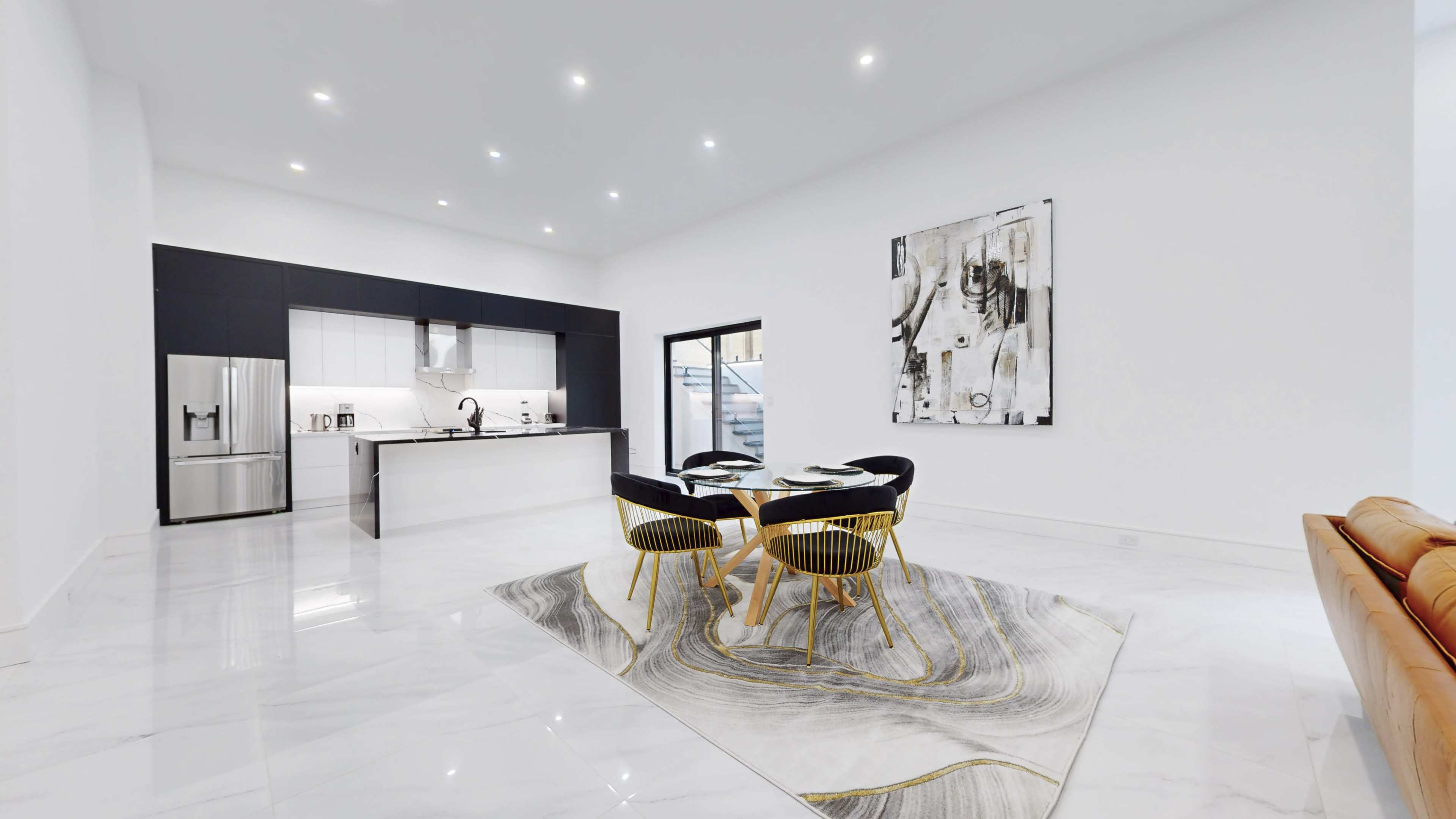 A modern kitchen and dining area featuring a large rug, a round dining table with four chairs, and a background of sleek cabinetry and appliances.
