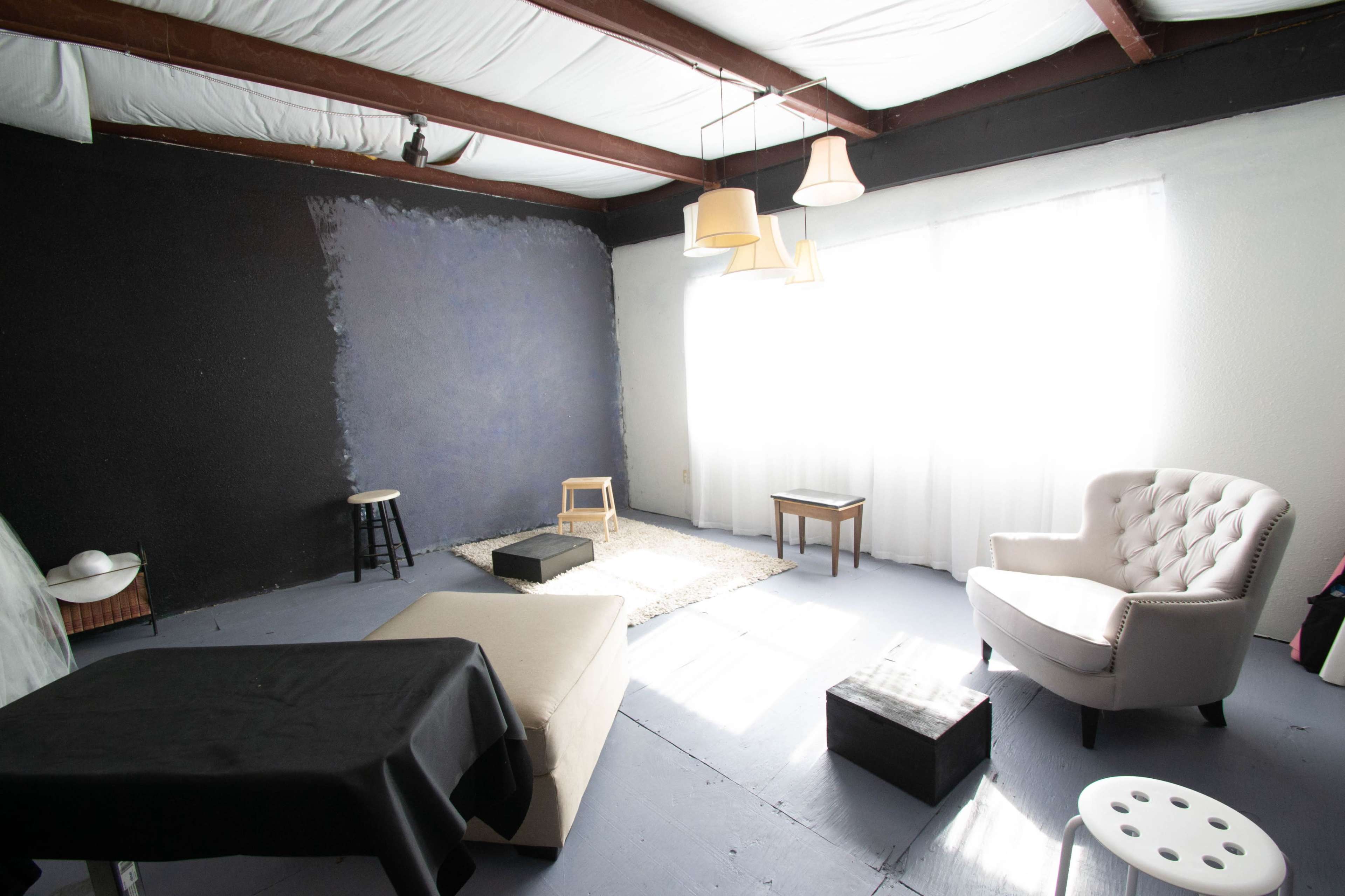 A sparsely furnished room with a mix of black and blue walls, a large window covered by sheer curtains, and a few simple pieces of furniture, including a white chair and a black table.