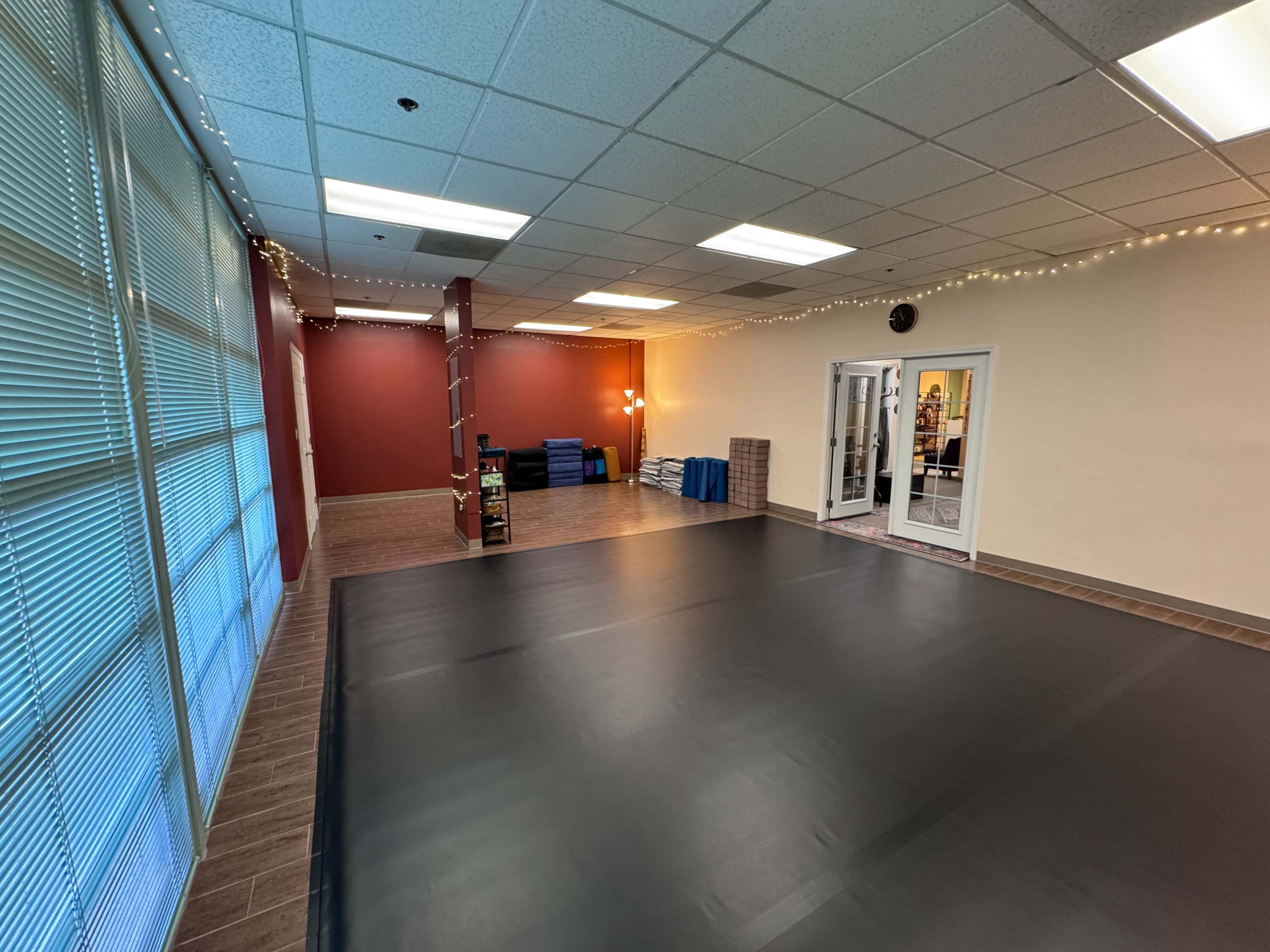 The image shows an empty room with a black mat flooring, large windows with blinds, and soft lighting along the ceiling.