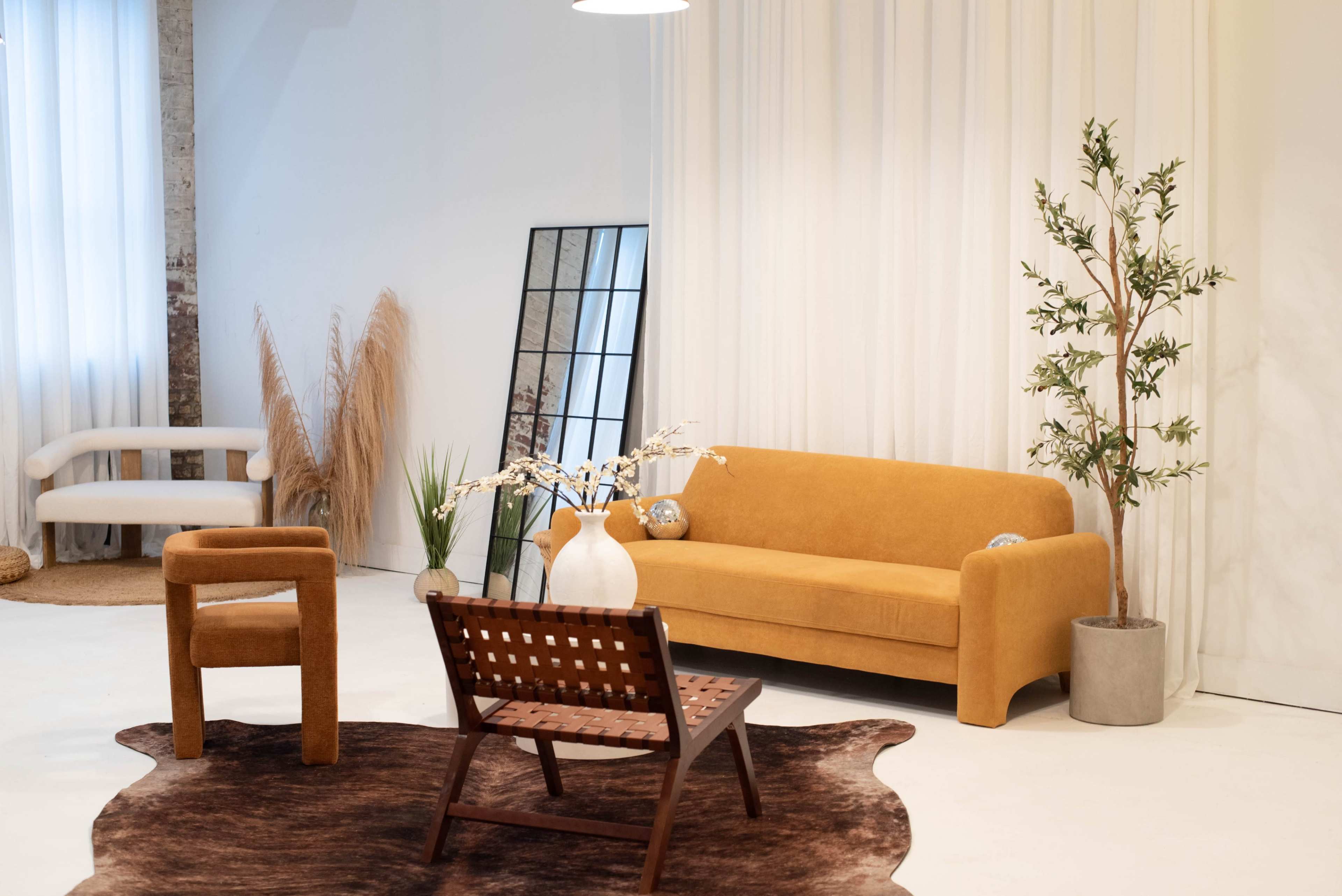 The image shows a bright, minimalist living space featuring a mustard yellow sofa, a brown lounge chair, a wooden slatted chair, decorative plants, and a mirror against a white wall.