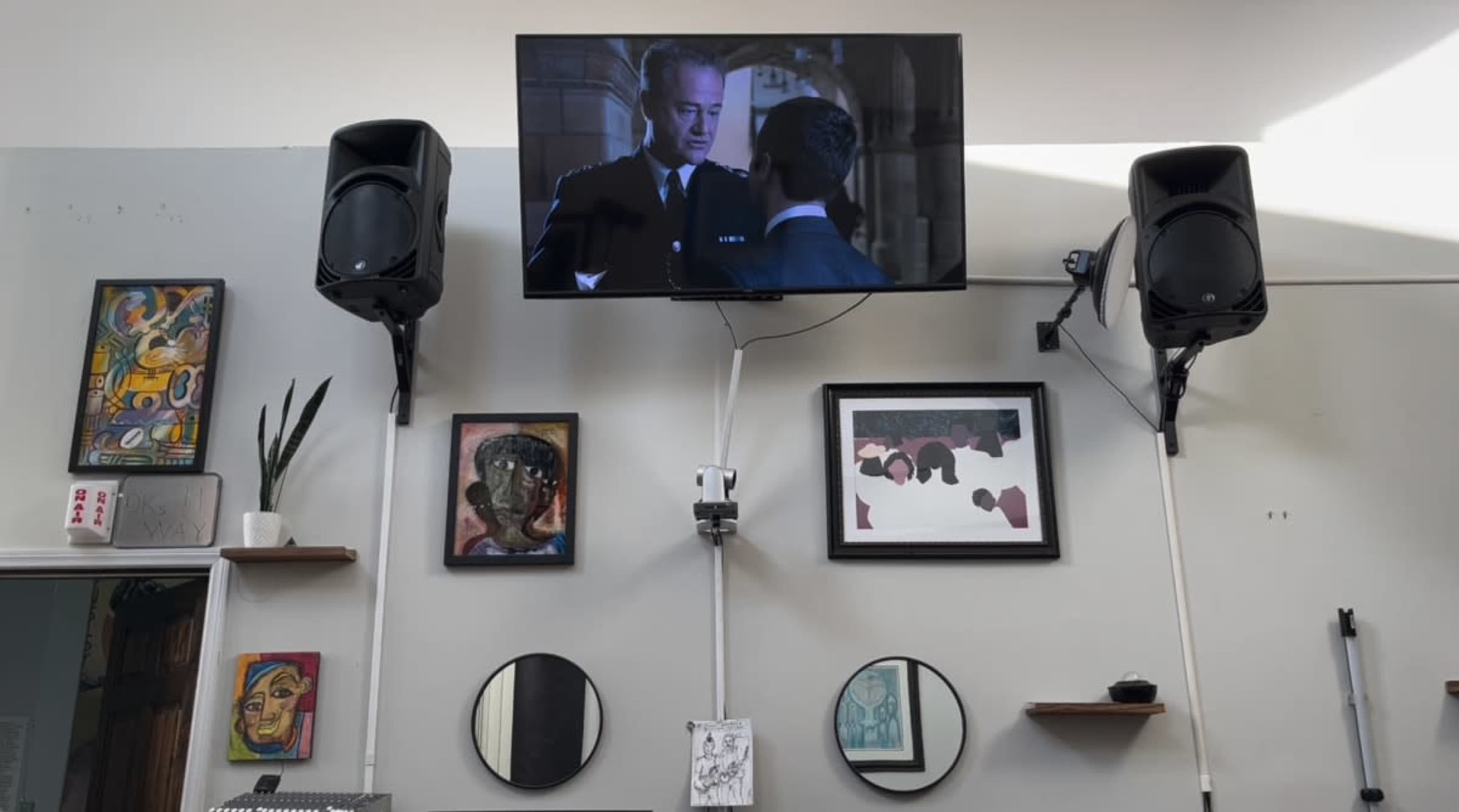 The image shows a wall with a mounted television displaying a scene from a movie, flanked by two large speakers, various pieces of artwork, and decorative mirrors.