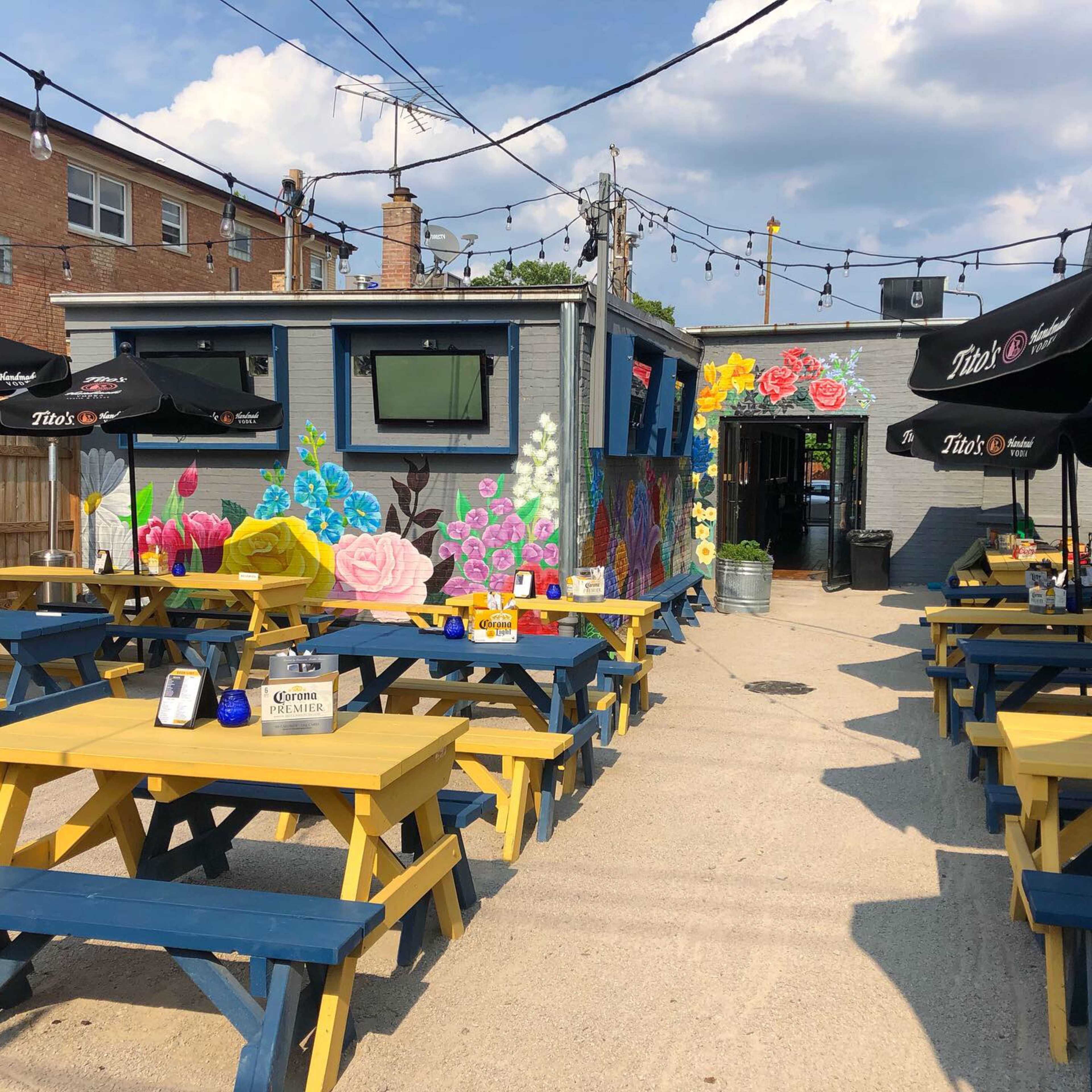 The image shows an outdoor seating area of a bar or restaurant featuring colorful picnic tables and a mural of flowers on the adjacent wall.