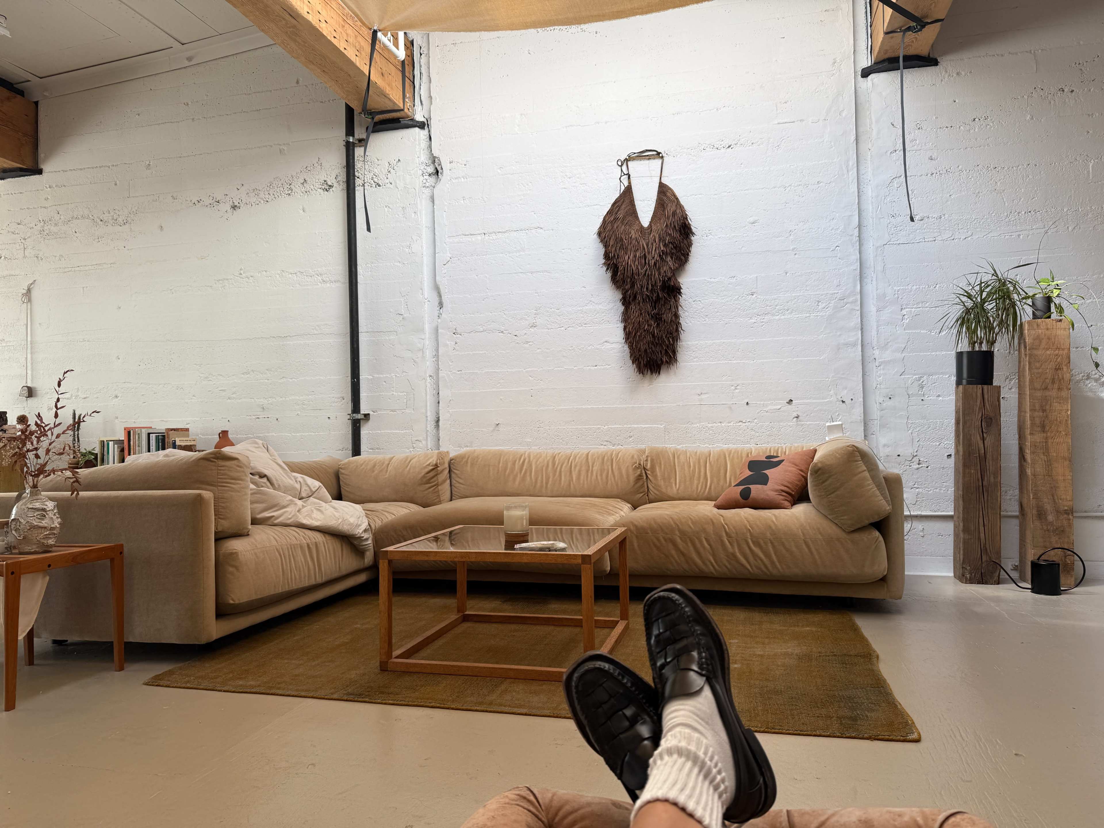 A modern living room features a light-colored sectional sofa, a glass coffee table on a beige rug, and a wall adorned with a decorative piece, framed by exposed beams.