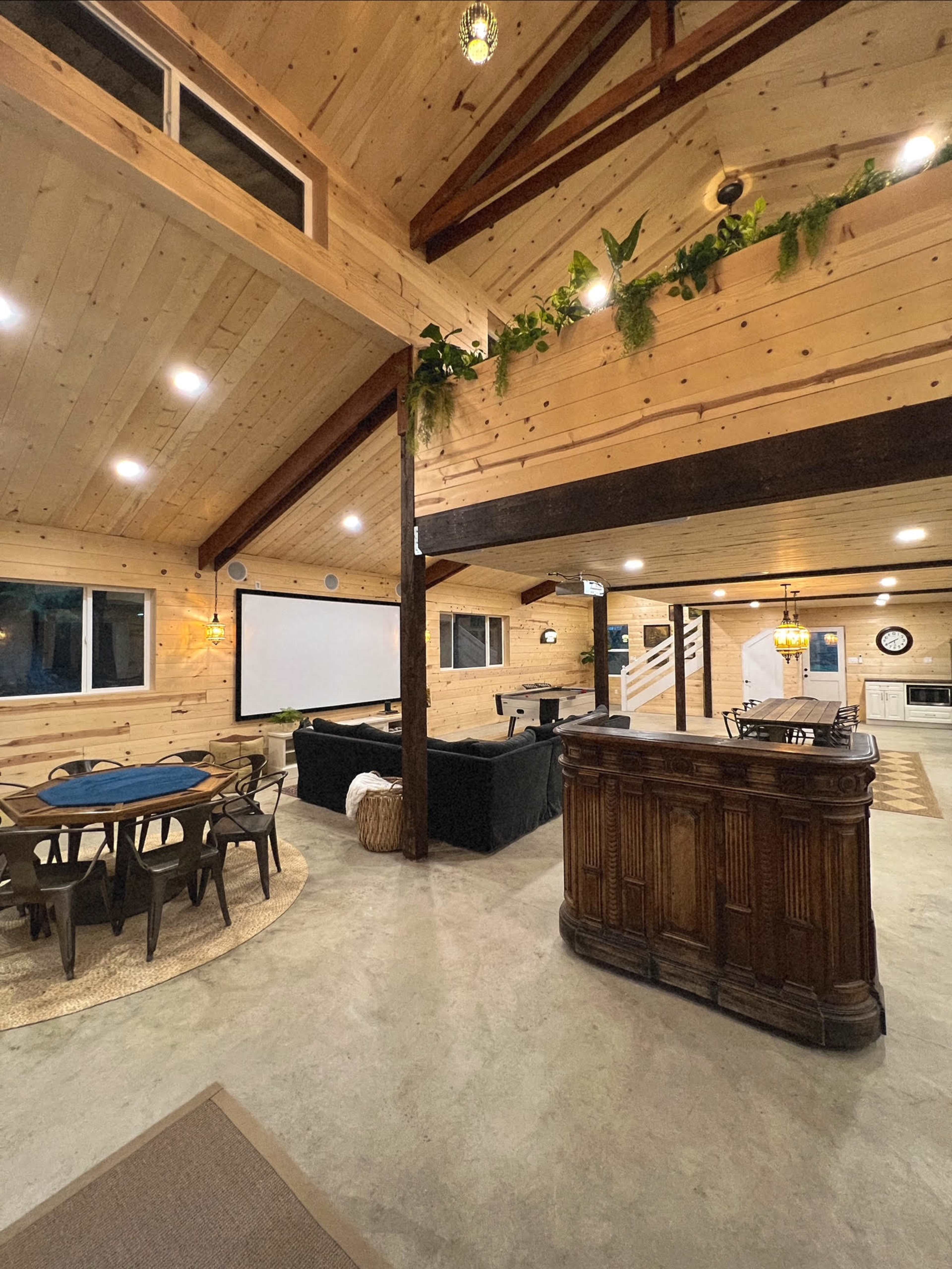 The image shows a spacious, wooden interior of a home featuring a dining area with a round table, a lounge area with a black sofa, and a bar counter, all illuminated by soft lighting.