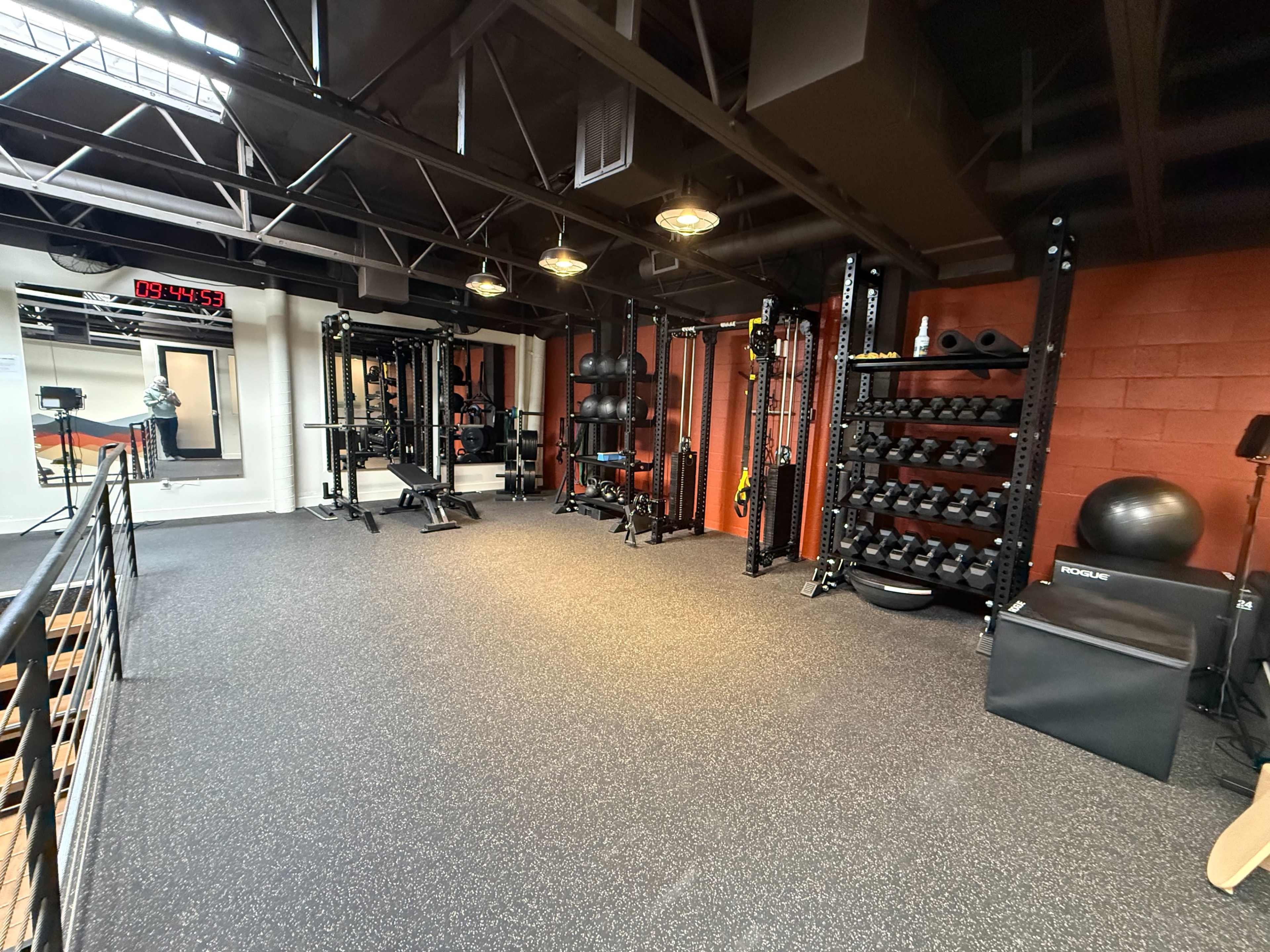 The image shows a well-equipped gym interior featuring weight racks, exercise equipment, and rubber flooring.