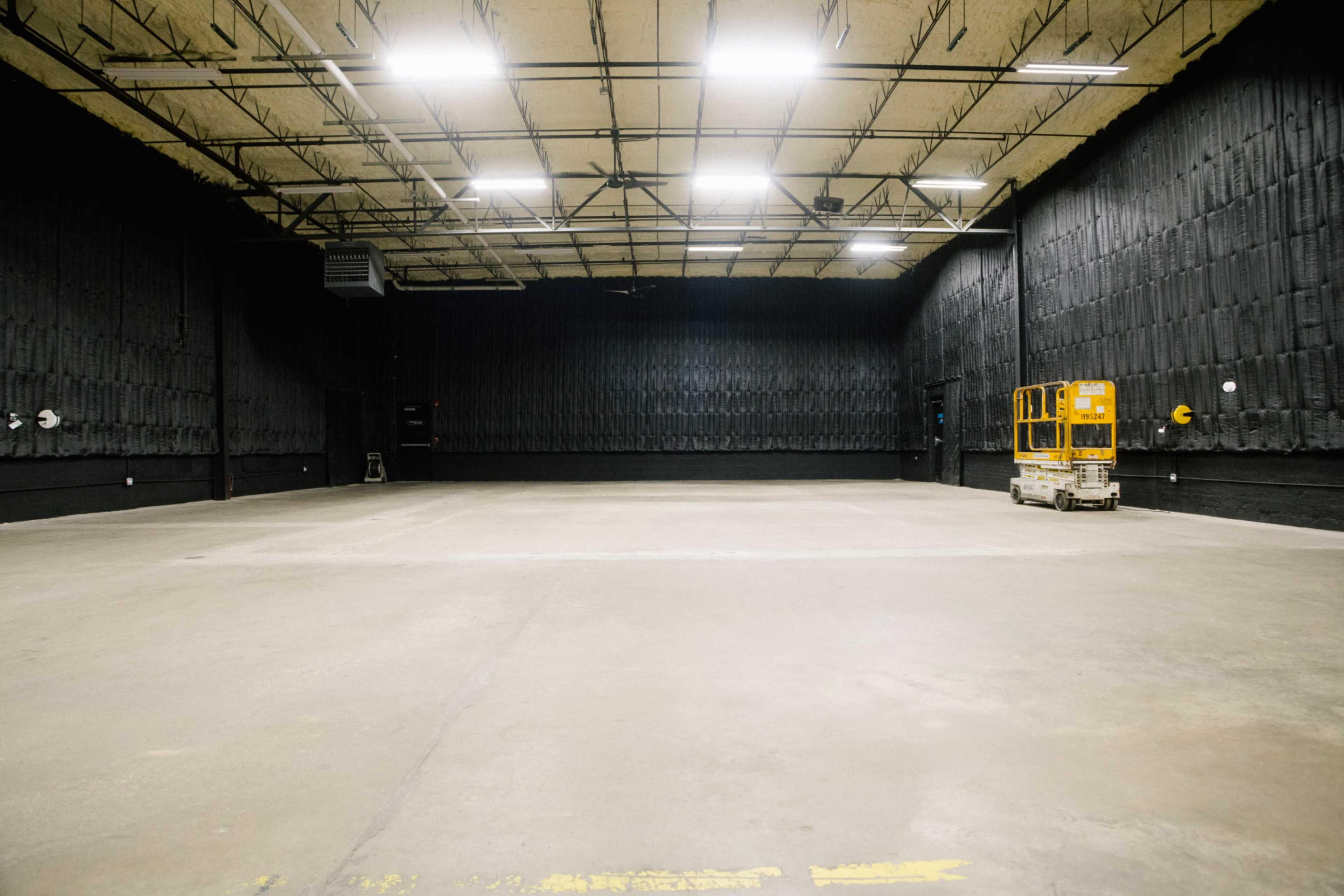 The image shows a large, empty industrial room with black textured walls and overhead lights, featuring a small yellow lift in one corner.