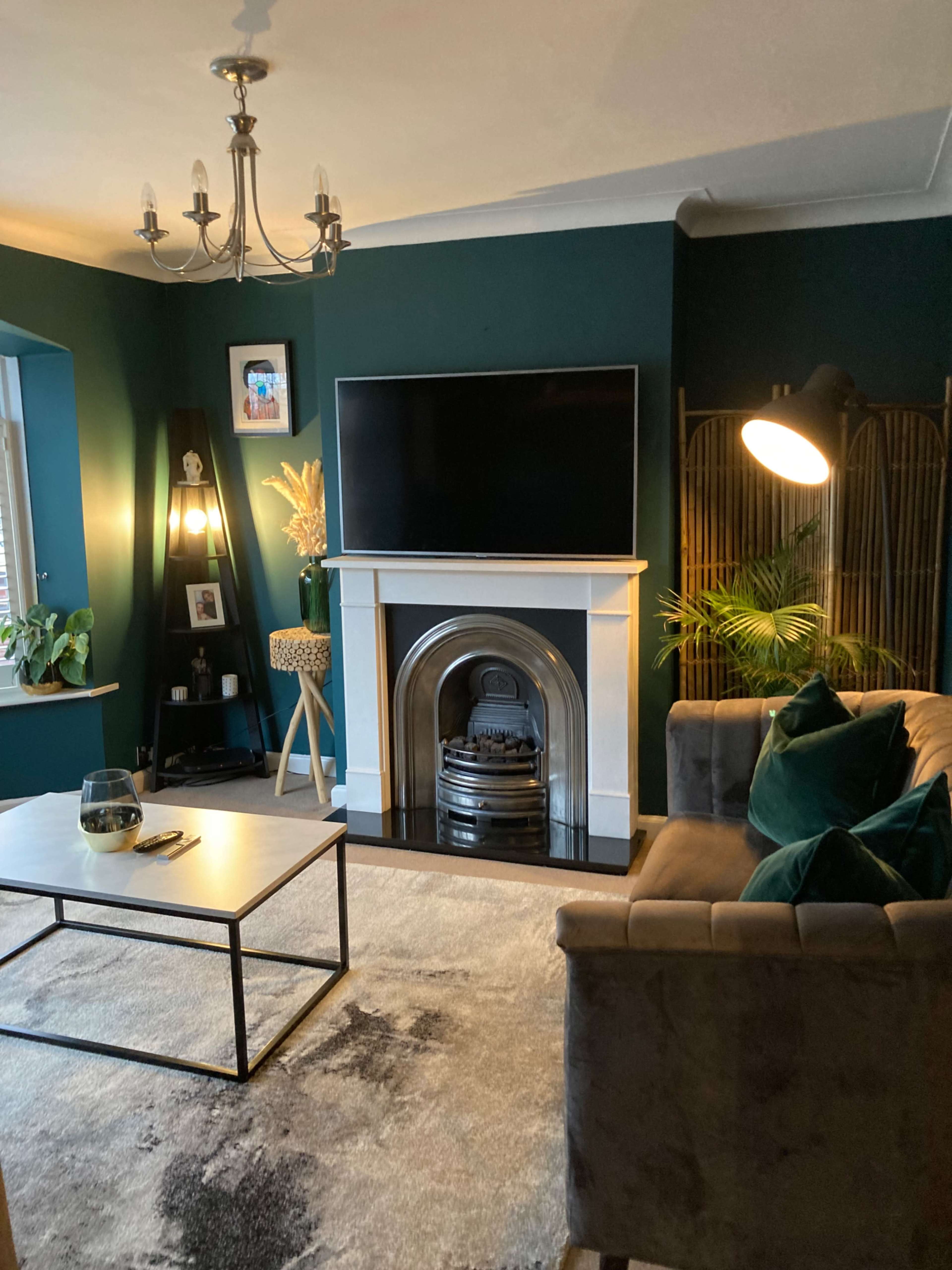 A cozy living room featuring a dark green wall, a large television above a white mantel, a stylish sofa, and a decorative lamp.
